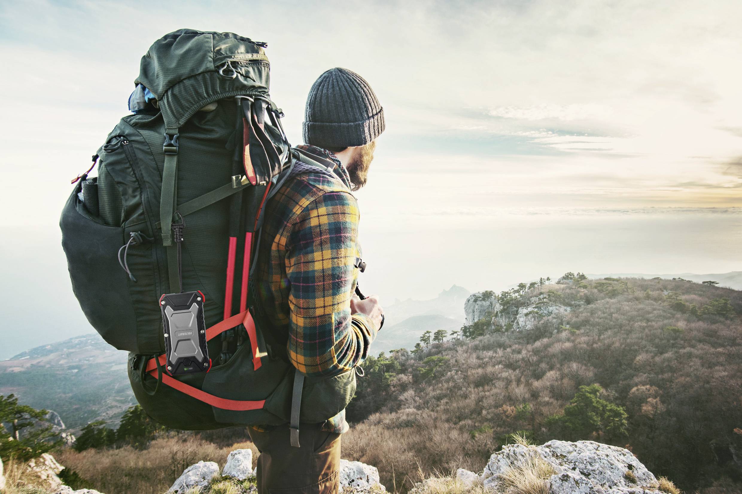 Person mit Rucksack und Mütze steht auf einem Aussichtspunkt und blickt über eine hügelige Landschaft in der Morgen- oder Abenddämmerung.
