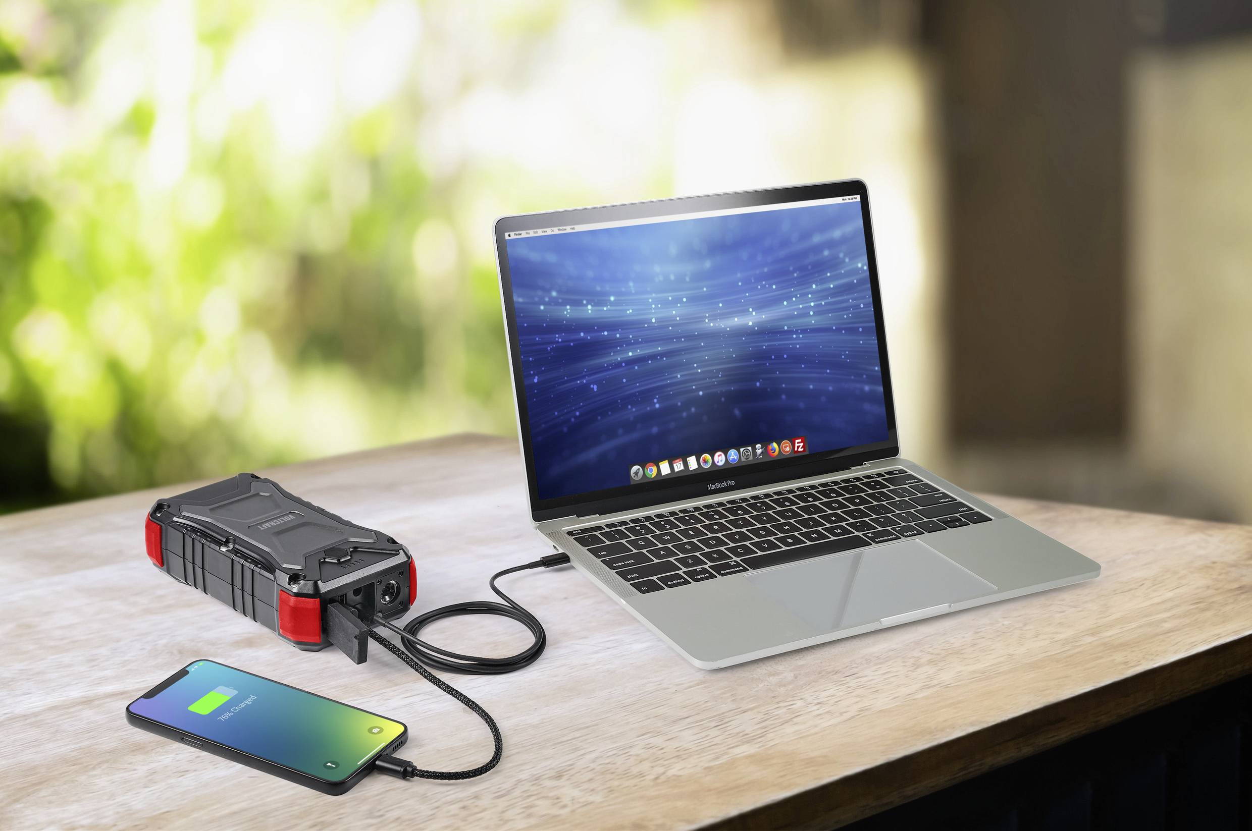 Ein Laptop und ein Smartphone laden über ein tragbares Ladegerät, das auf einem Holztisch steht. Im Hintergrund sind unscharfe Pflanzen erkennbar.