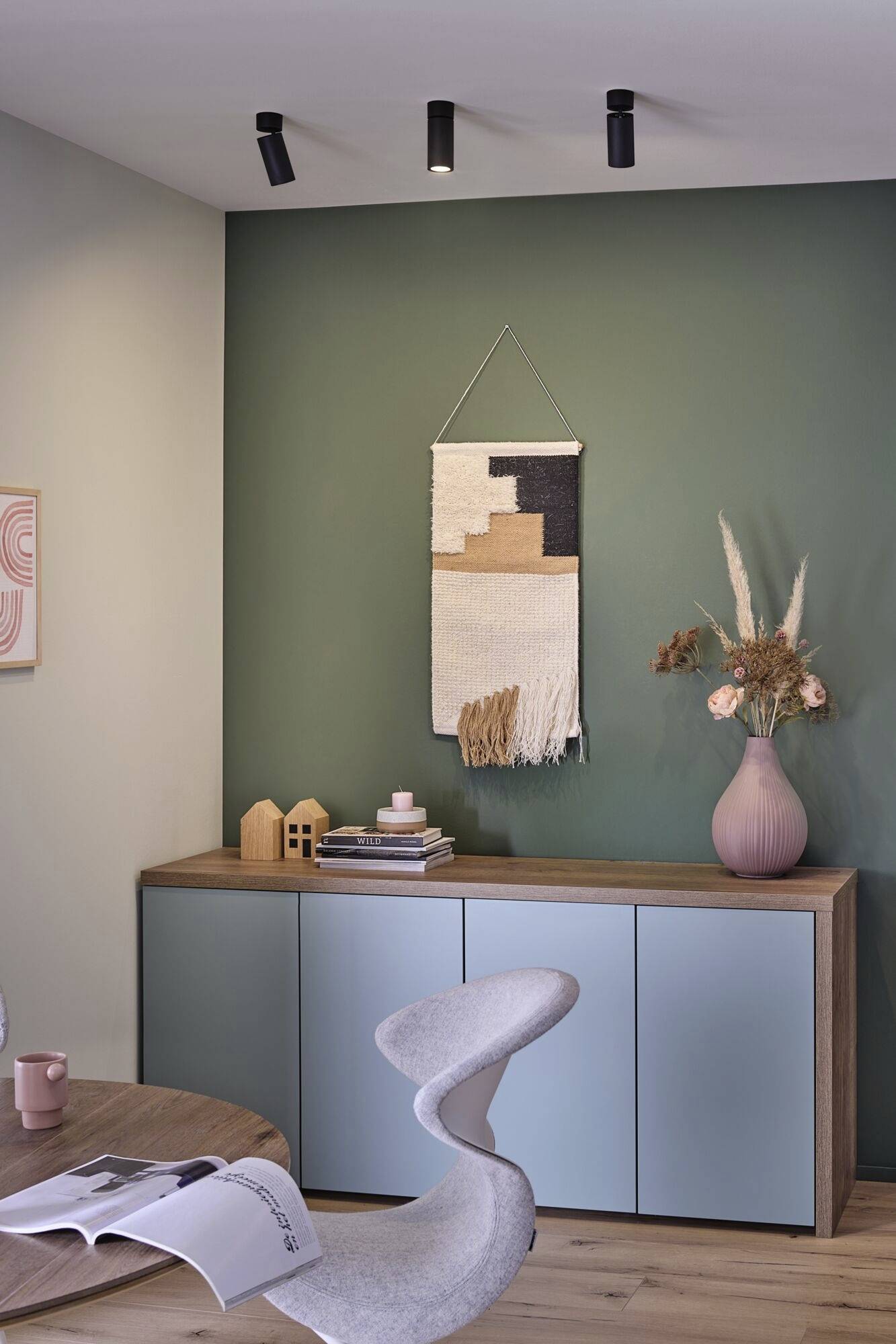 Ein modernes Wohnzimmer mit eleganten Möbeln. Ein grünes Wandbild, Holzregal mit Dekorationen und ein geschwungener Stuhl sind sichtbar.