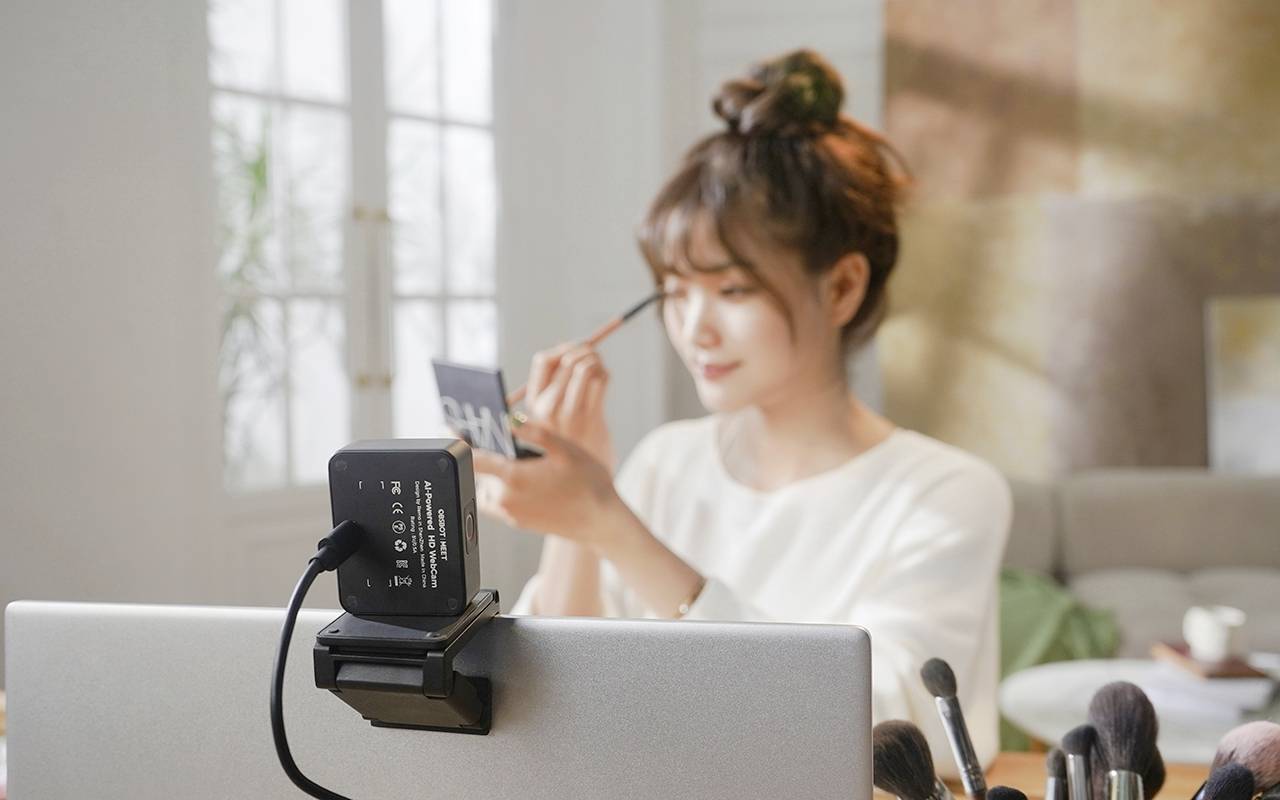 Eine Person sitzt vor einem Computer und trägt mit einem Pinsel Make-up auf ihr Gesicht auf. Im Vordergrund ist eine Kamera zu sehen.