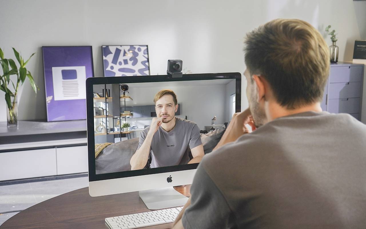 Ein Mann sitzt an einem Tisch und schaut auf einen Computerbildschirm, auf dem ein anderer Mann in einem Videoanruf zu sehen ist.