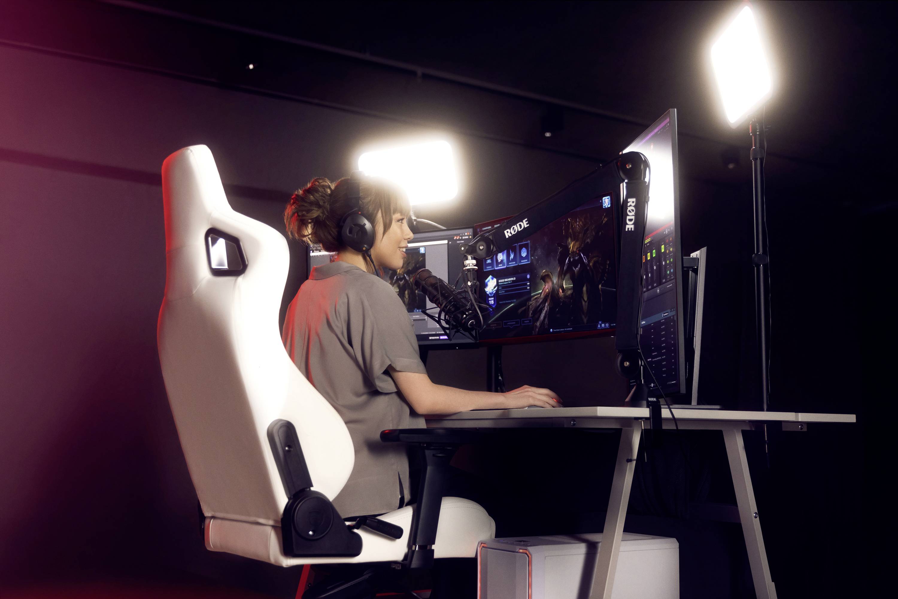 Eine Person mit Kopfhörern sitzt an einem Gaming-Schreibtisch und spielt ein Videospiel. Zwei Bildschirme und Lichter im Hintergrund.