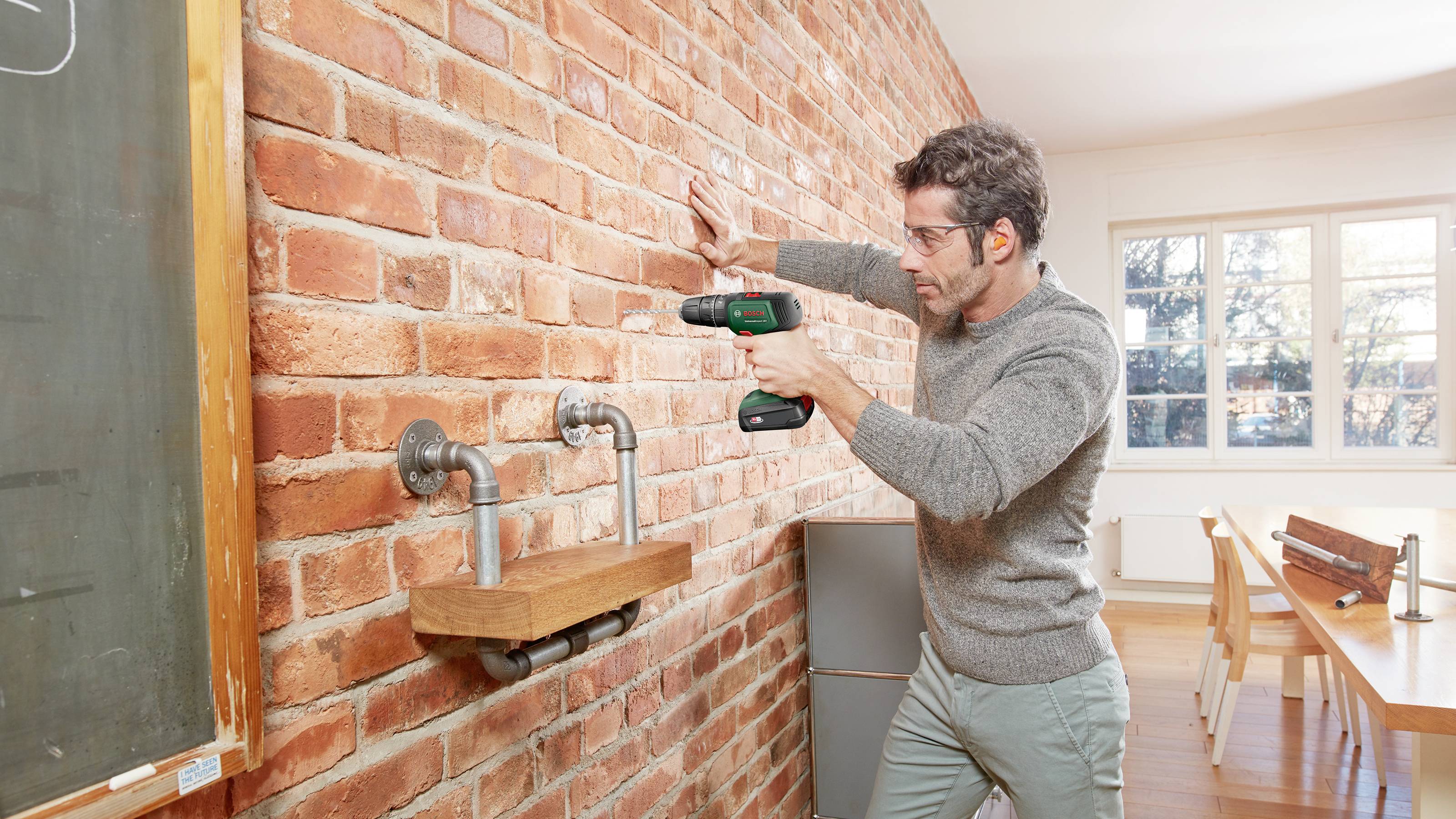 Ein Mann in einem Raum befestigt mit einer Bohrmaschine ein Regal an einer Backsteinwand. Im Hintergrund sind Fenster und Möbel.
