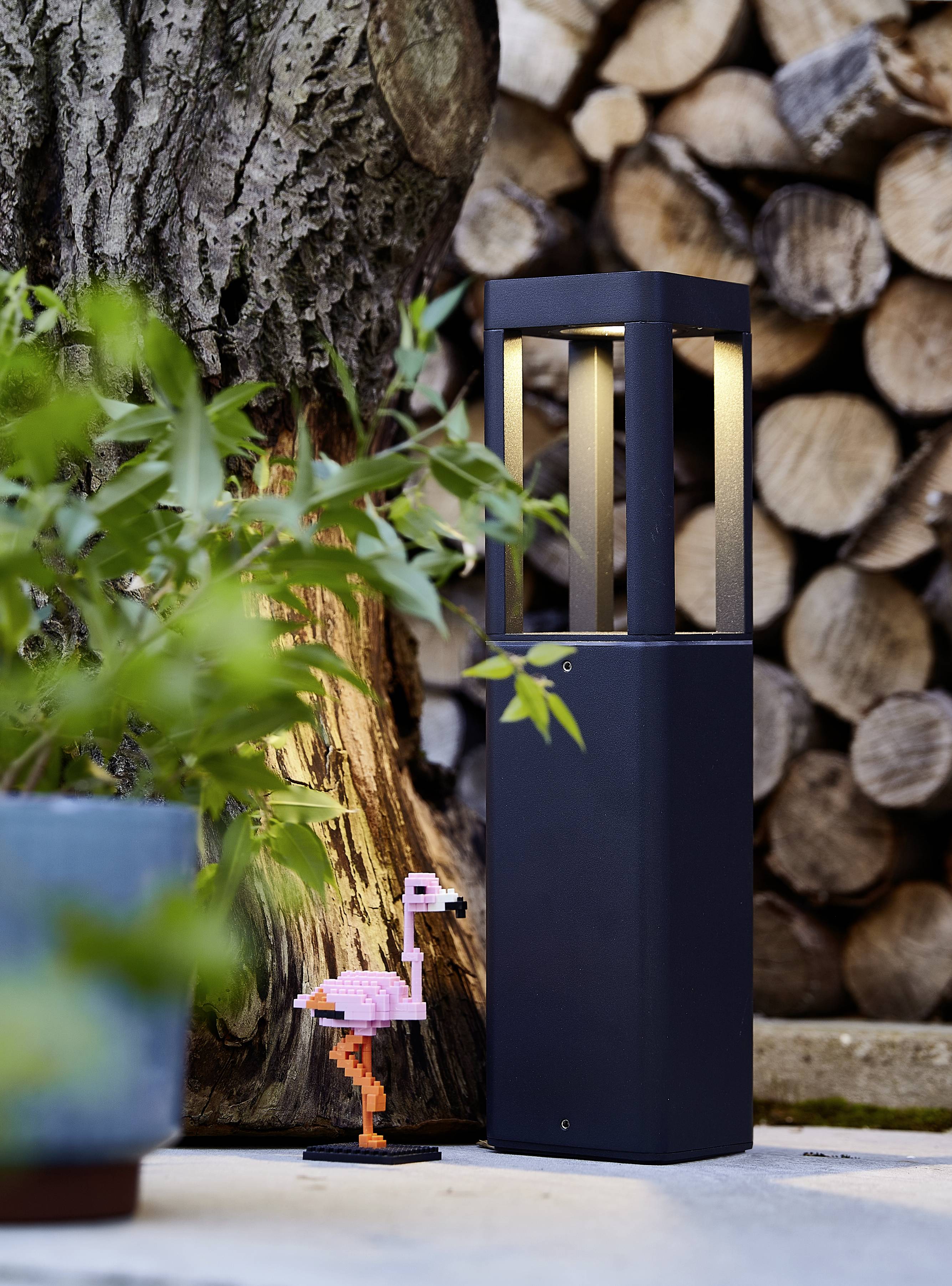 Außenstehende schwarze Lampe neben einem Baumstamm und Holzstapel, beleuchtet einen kleinen, rosa Flamingo-Dekorationsgegenstand.