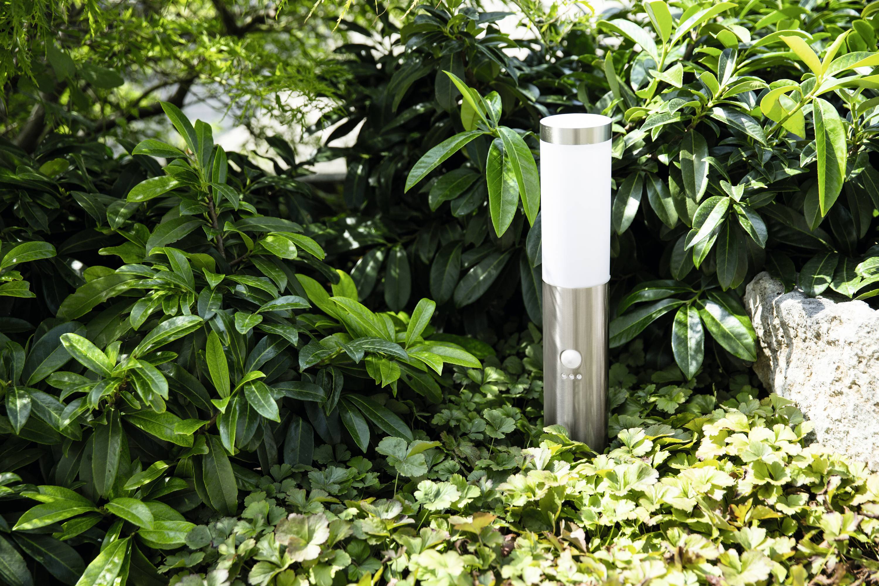 Eine moderne, silberne Außenleuchte steht zwischen üppigen, grünen Pflanzen im Garten und erzeugt einen Kontrast zu den Blättern.