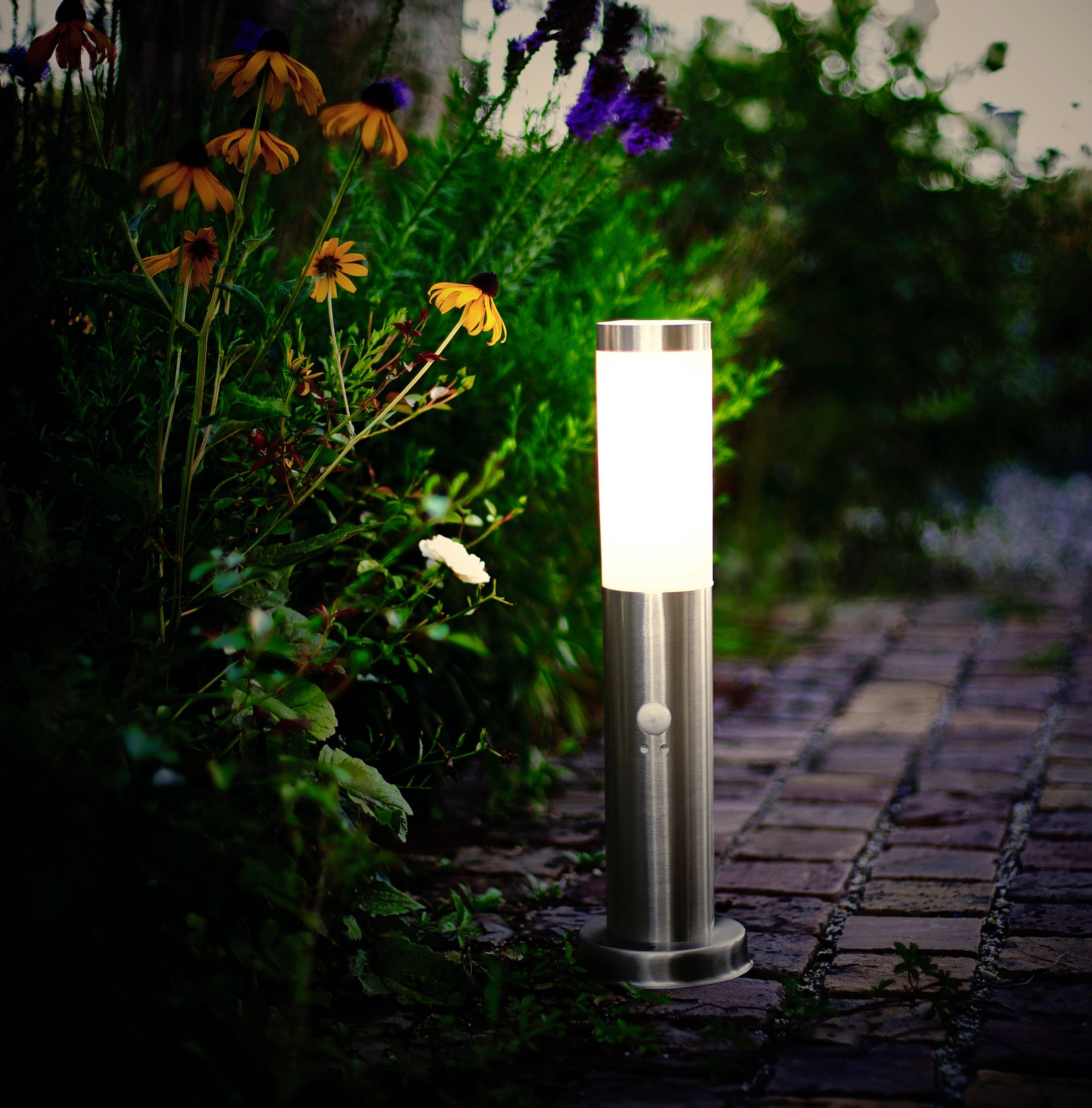 Eine leuchtende Gartenlampe steht neben einem Weg aus Kopfsteinpflaster, umgeben von Blumen und Pflanzen in der Dämmerung.