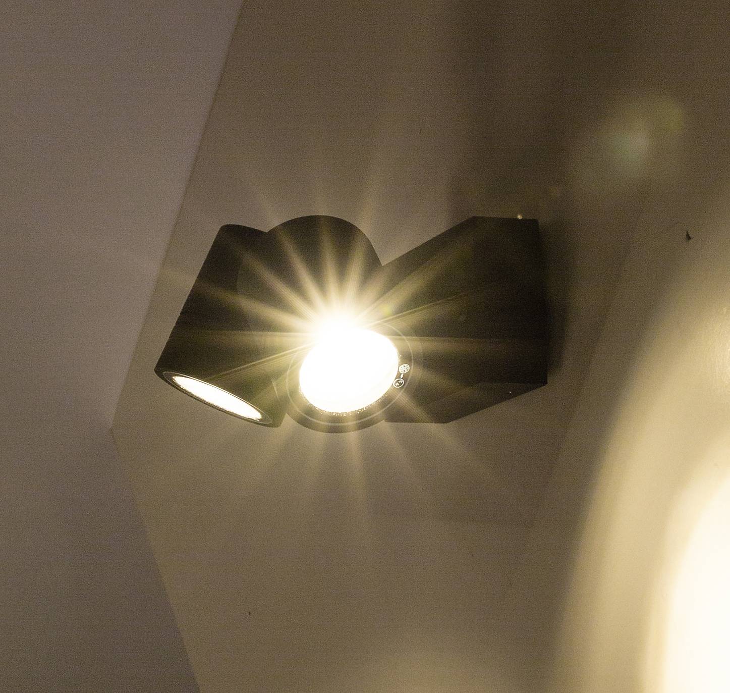 Wandleuchte mit zwei Strahlern an, einer leuchtet hell. Beleuchtet eine Wand.