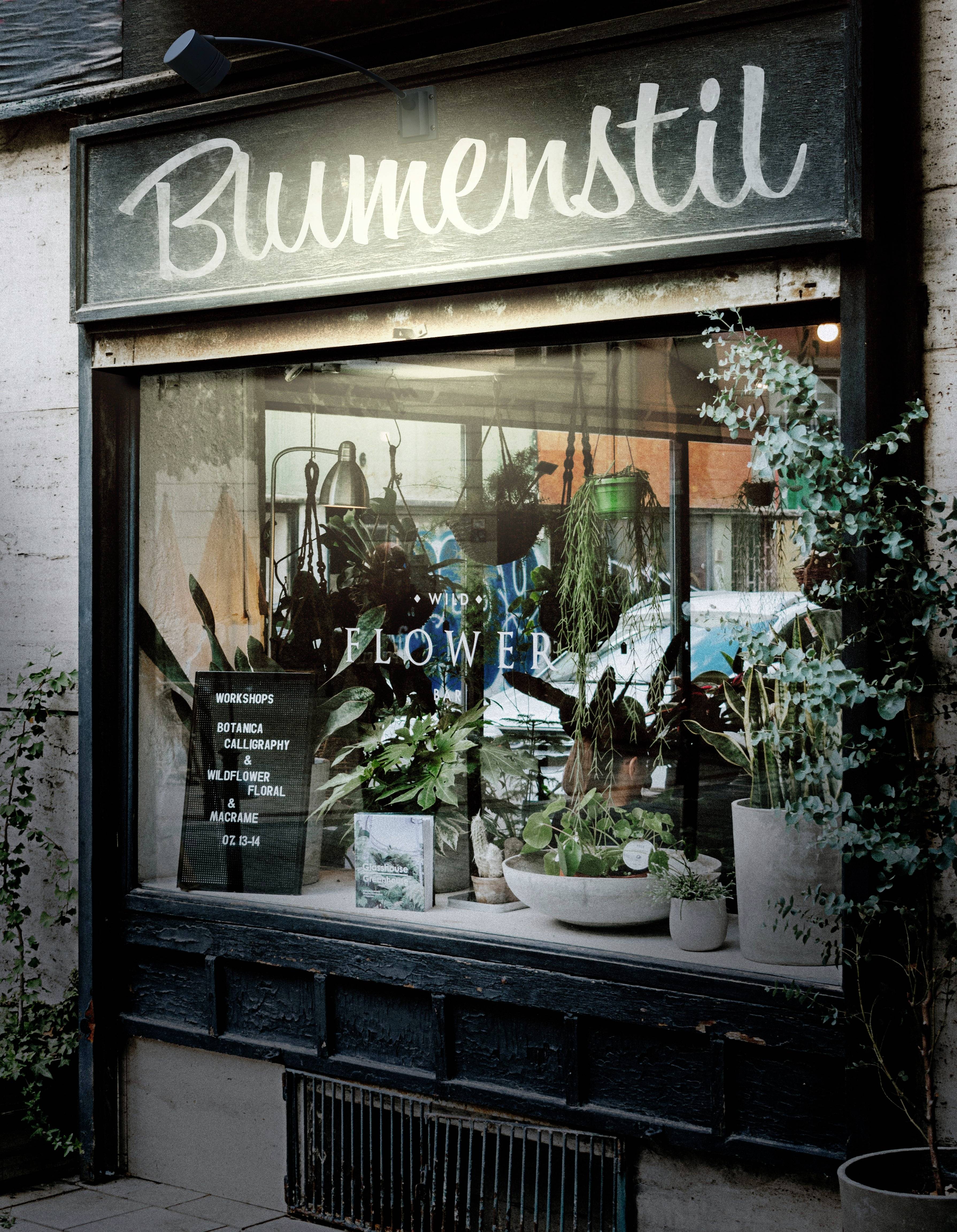 Ein Schaufenster eines Blumengeschäfts mit der Aufschrift 'Blumenstil' und verschiedenen Pflanzen in Töpfen ausgestellt.