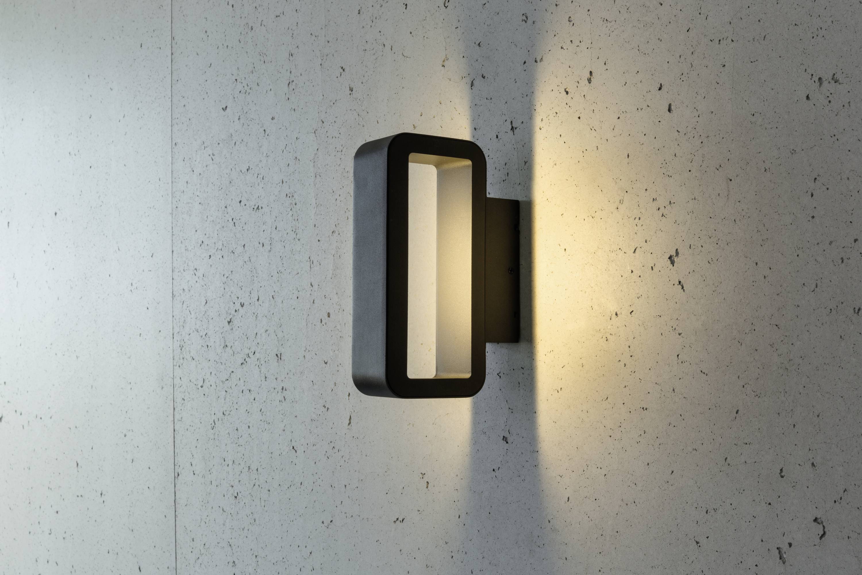 Wandleuchte mit rechteckigem Rahmen, leuchtet warmes Licht auf eine strukturierte Wand. Modernes, minimalistisches Design.