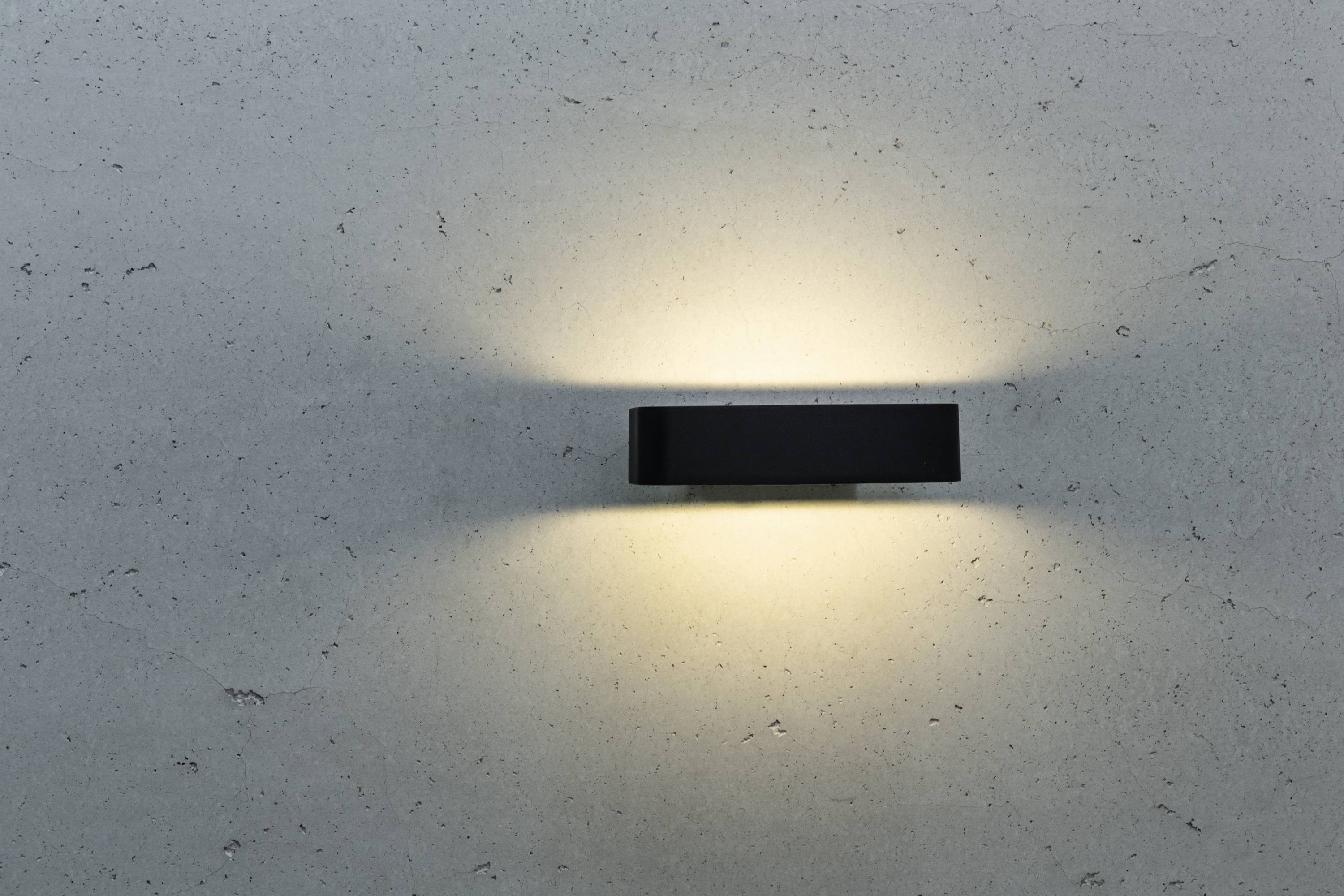 Wandleuchte an einer grauen Wand, die sanftes, diffuses Licht nach oben und unten ausstrahlt, erzeugt eine gemütliche Atmosphäre.