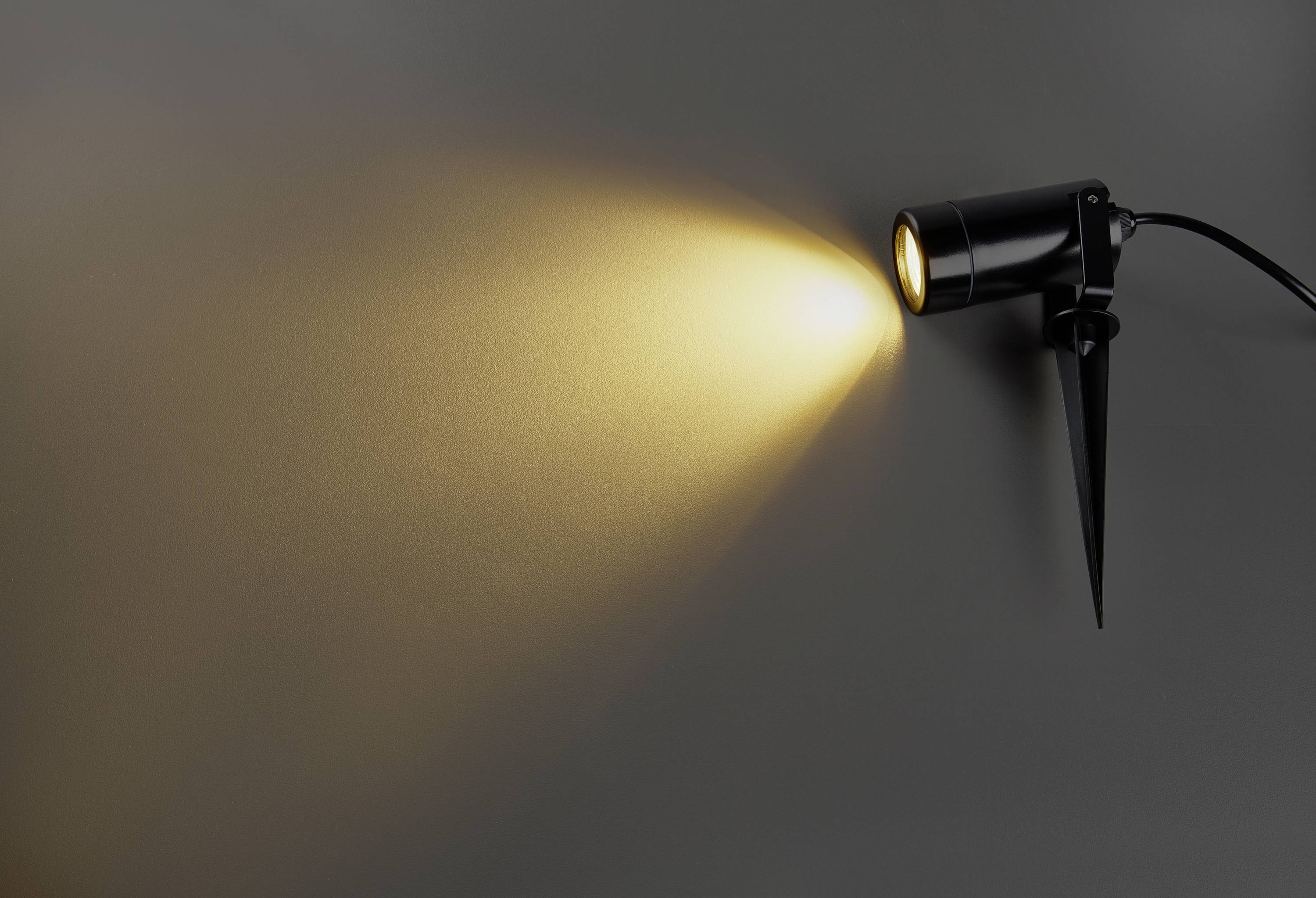 Ein schwarzer Strahler an der Wand wirft einen warmen Lichtkegel nach rechts auf die graue Fläche.