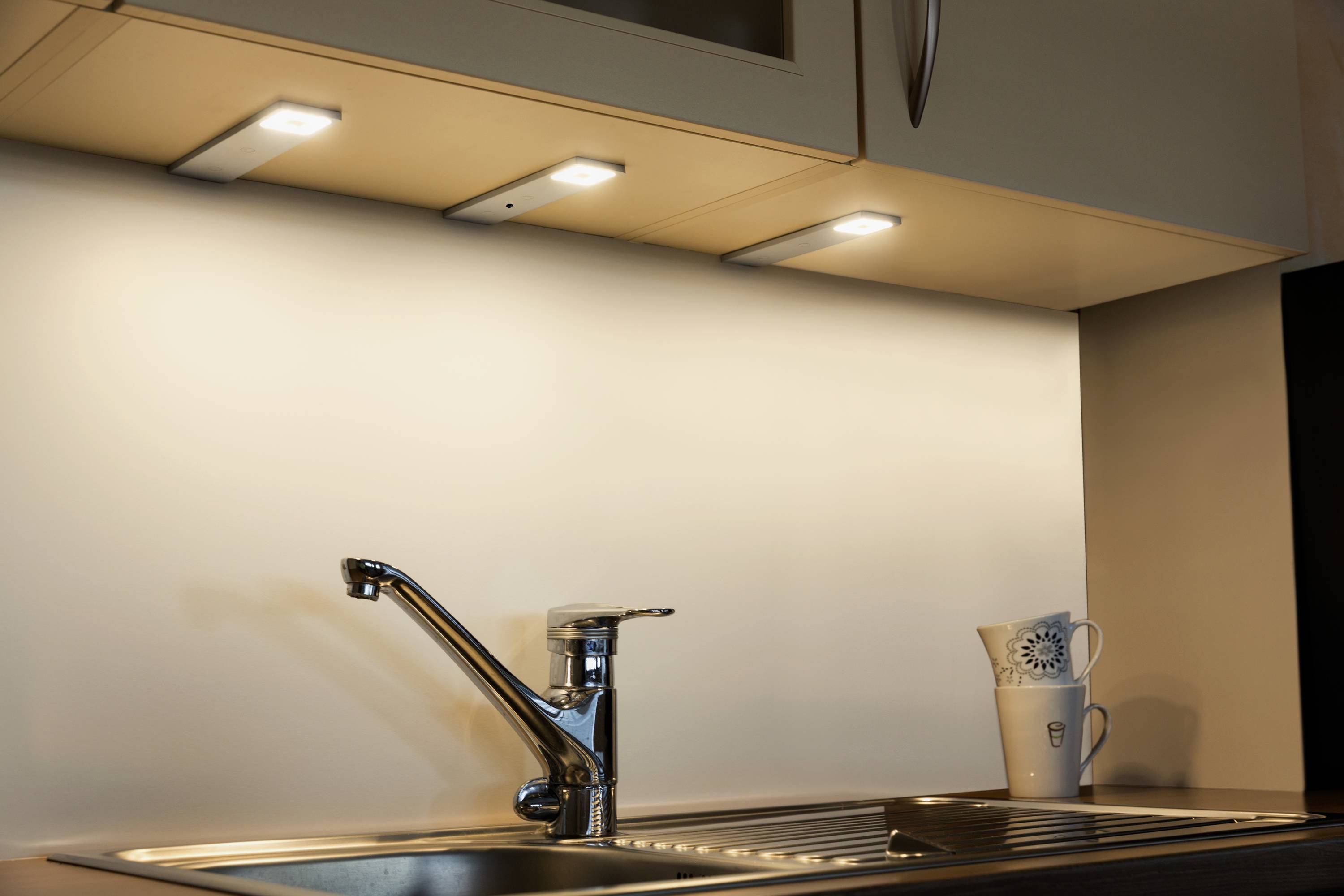 Eine Küche mit Edelstahlarmatur und Spüle. Unter den Hängeschränken sind drei LED-Leuchten angebracht, die die Arbeitsfläche beleuchten. Im Hintergrund eine dekorative Tasse.
