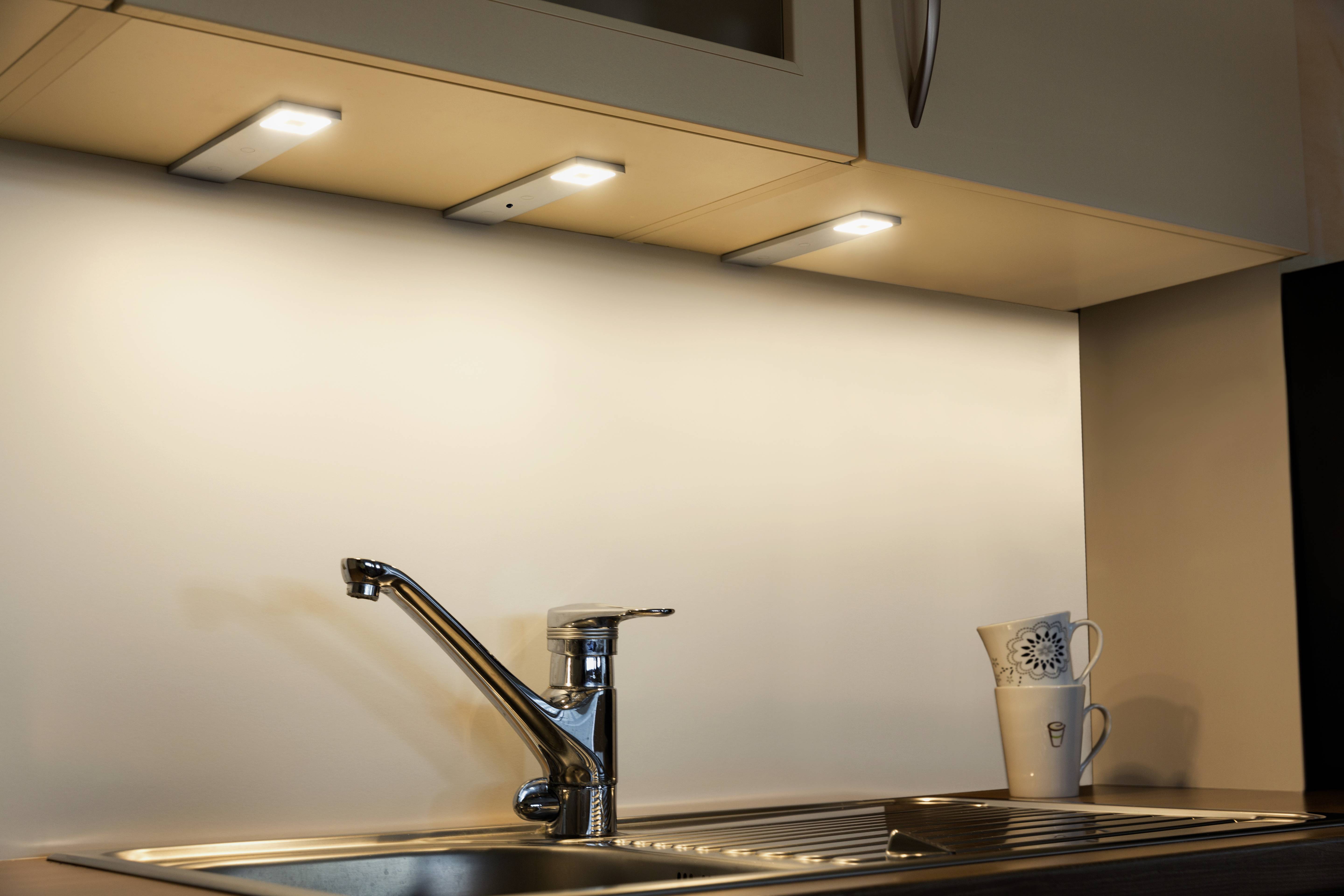 Küchenarbeitsfläche mit integriertem Waschbecken und Wasserhahn. Drei LED-Leuchten unter den Schränken beleuchten die Arbeitsfläche. Eine Tasse mit Sonnenblumenmotiv steht am Rand.