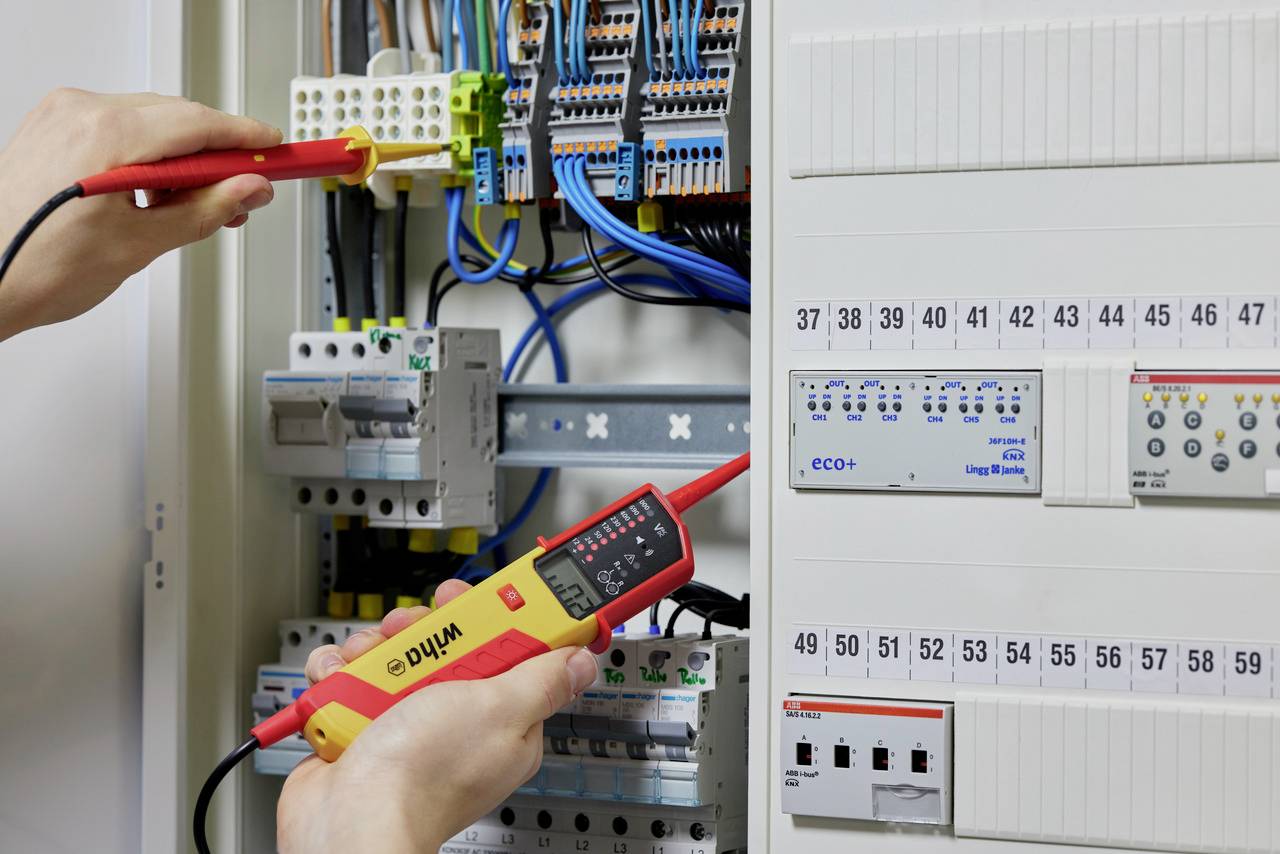 Eine Person testet mit einem Multimeter die elektrischen Verbindungen in einem geöffneten Schaltschrank voller Kabel und Sicherungen.