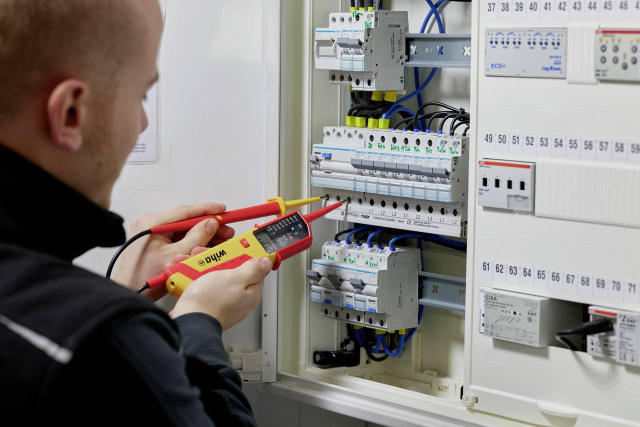 Eine Person prüft mit einem Multimeter die elektrischen Anschlüsse in einem geöffneten Sicherungskasten.
