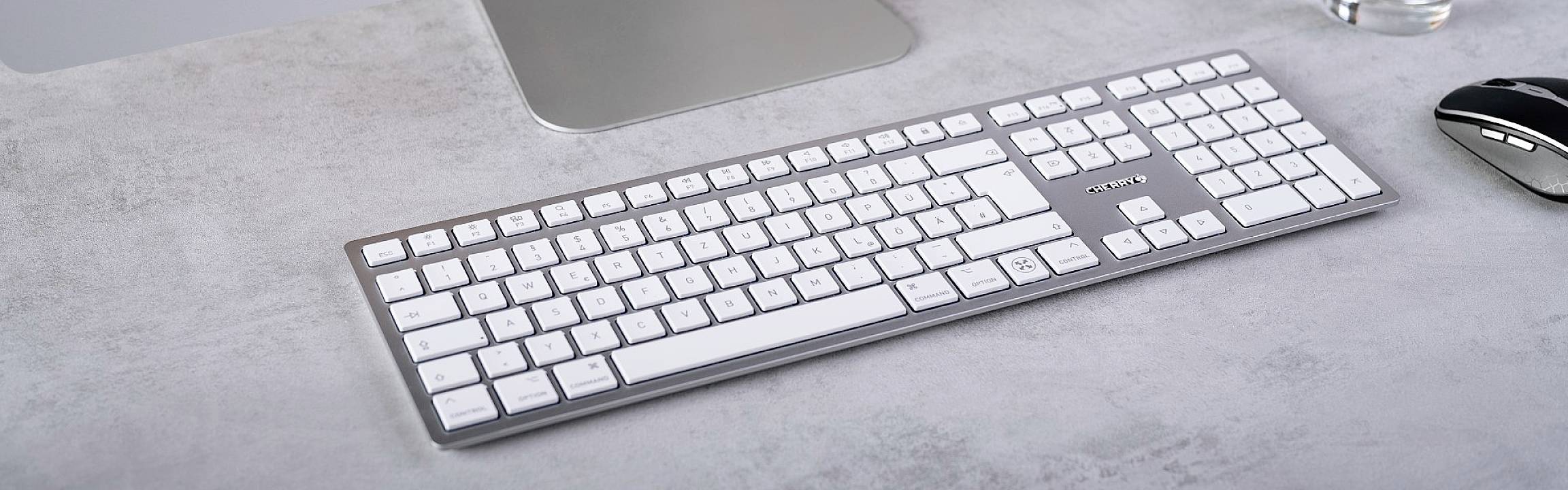 Weiße Computertastatur auf einem grauen Schreibtisch neben einer Computermaus, mit einem Teil eines Bildschirms sichtbar.