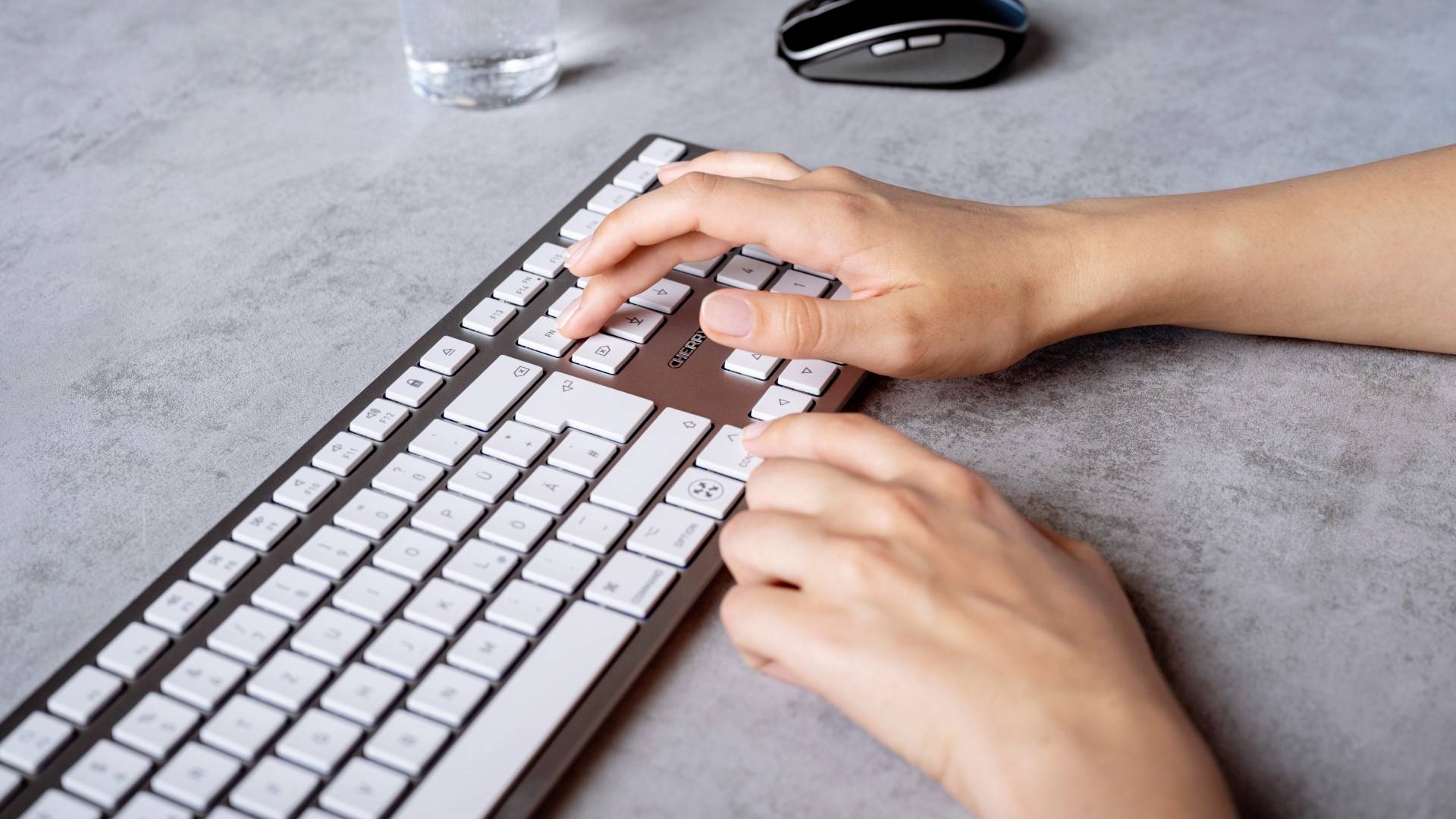 Hände tippen auf einer Computertastatur auf einem grauen Schreibtisch. Im Hintergrund sind eine Maus und ein Glas Wasser zu sehen.