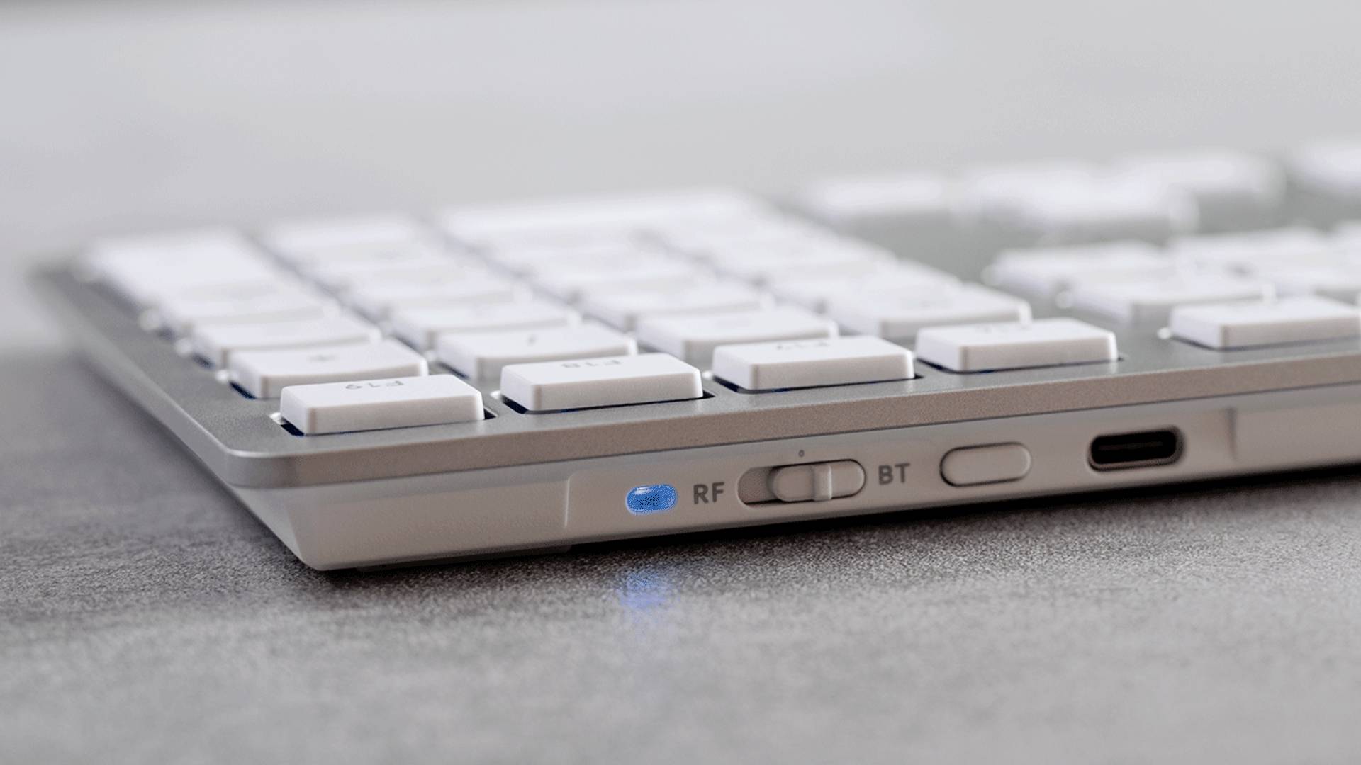 Eine drahtlose Tastatur mit aktivierter Bluetooth-Funktion, erkennbar am blauen Licht. Tasten in Nahaufnahme, grauer Hintergrund.