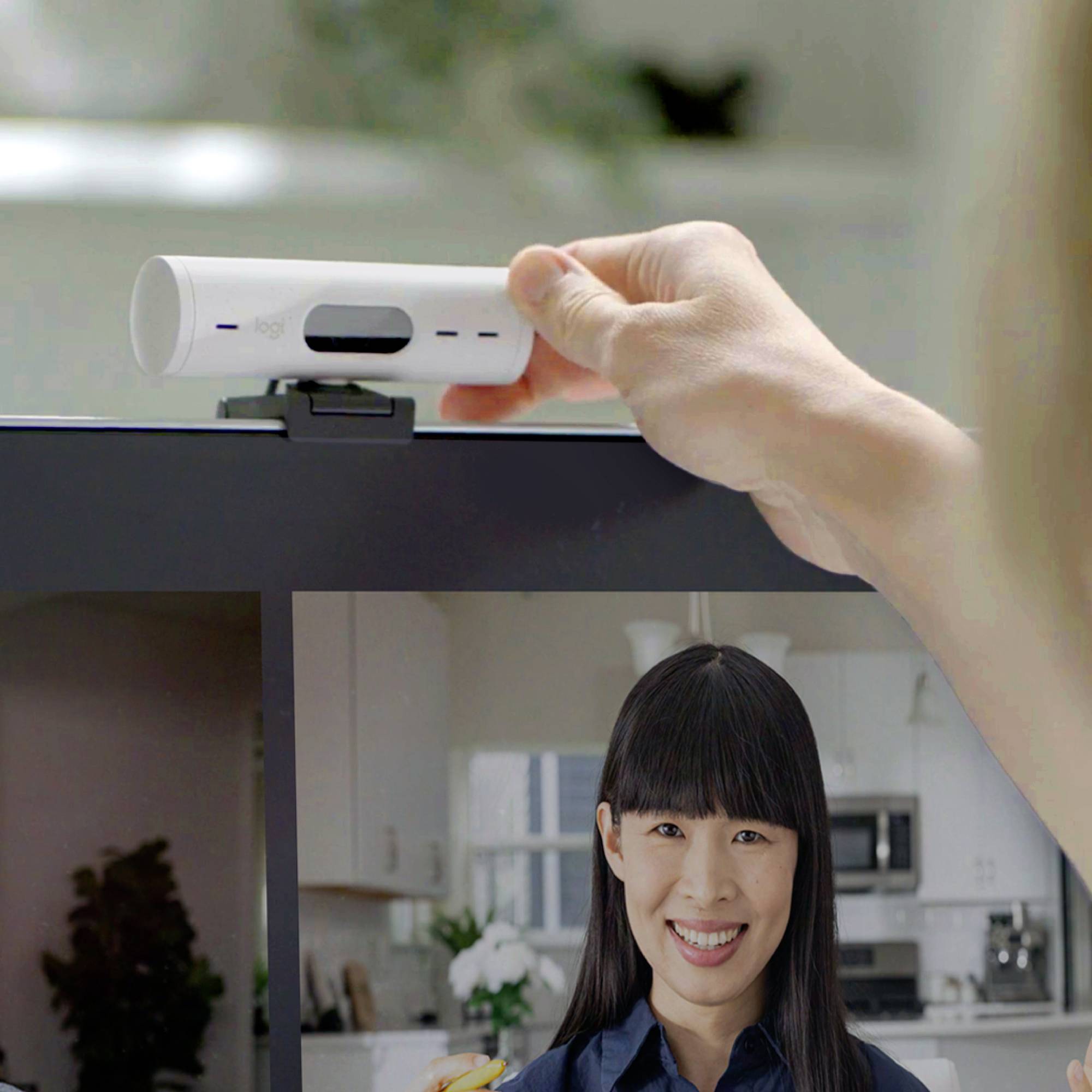 Eine Person lächelt in einem Videocall, während eine Hand eine Webcam auf dem Computerbildschirm anpasst. Im Hintergrund ist eine Küche erkennbar.