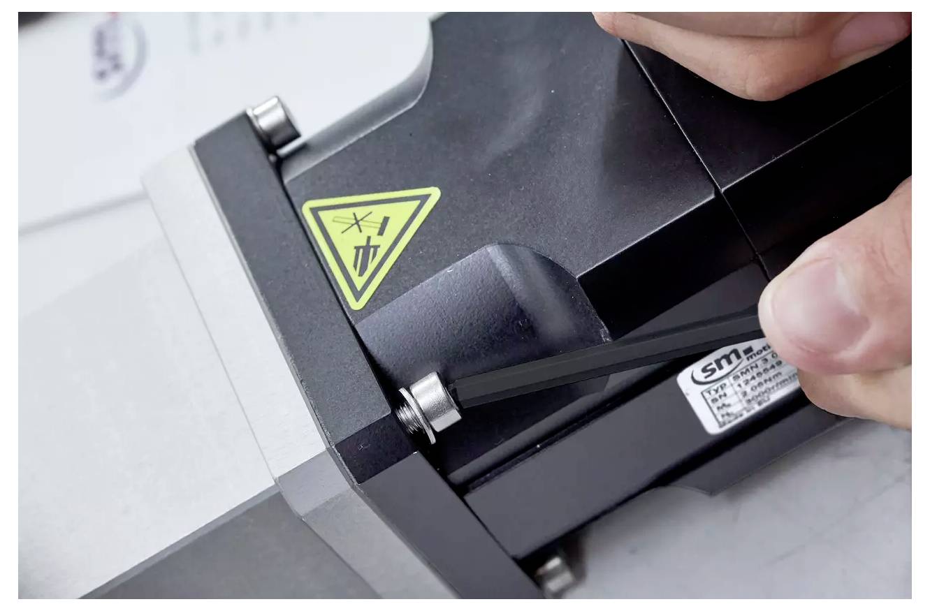 Eine Hand, die ein Werkzeug an einem Metallgerät mit einem gelben dreieckigen Sicherheitswarnschild verwendet, das auf eine mechanische Gefahr hinweist.