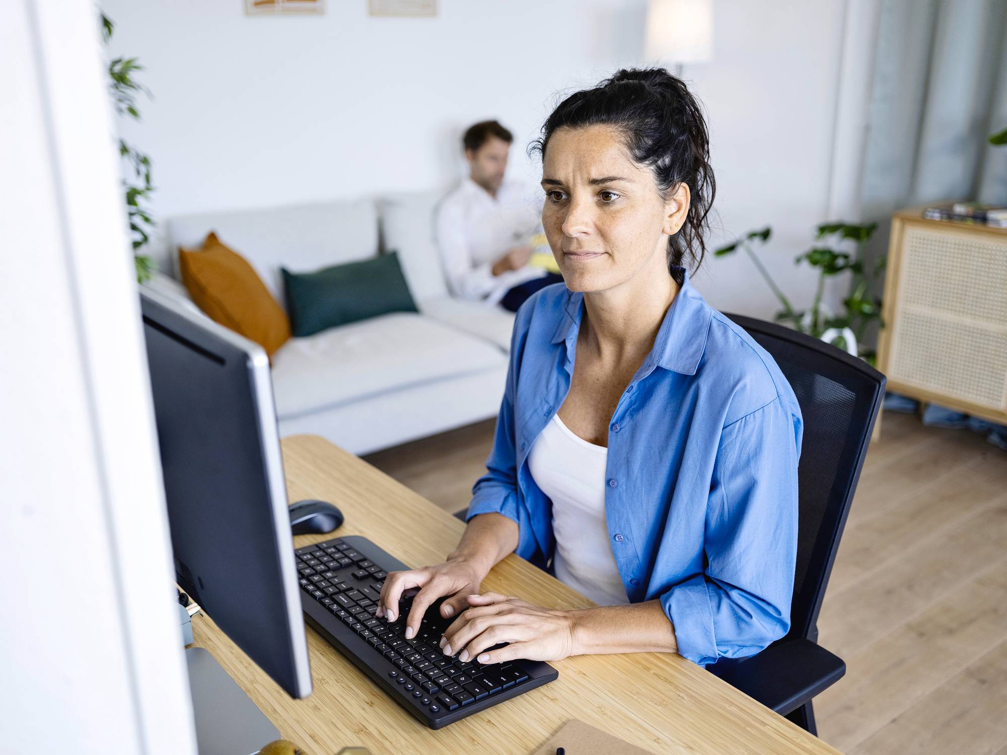 Frau am Schreibtisch arbeitet am Computer, während ein Mann im Hintergrund auf einem Sofa sitzt. Wohnzimmerumgebung.