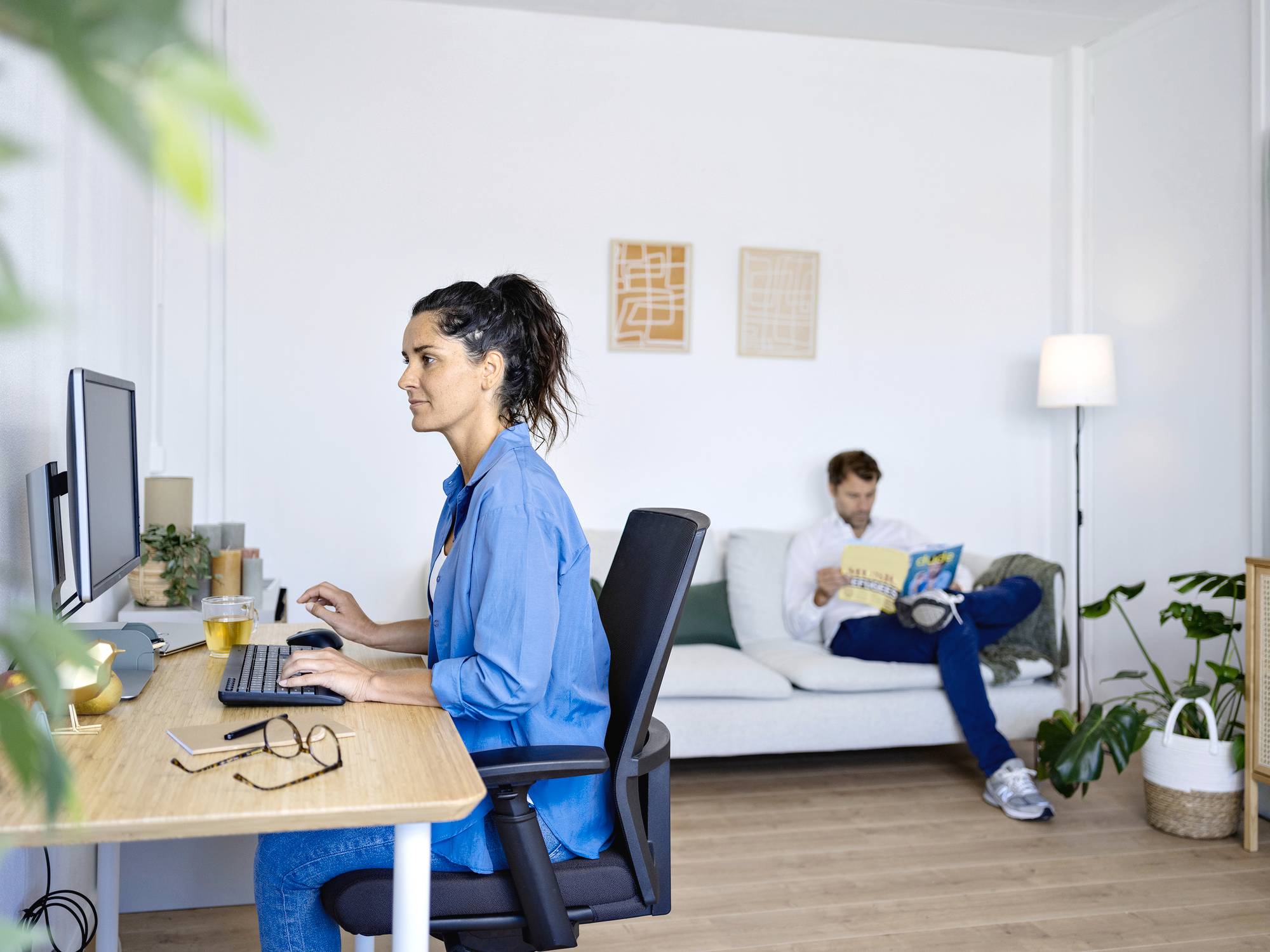 Eine Frau arbeitet am Schreibtisch am Computer, während ein Mann im Hintergrund auf einem Sofa sitzt und ein Buch liest.