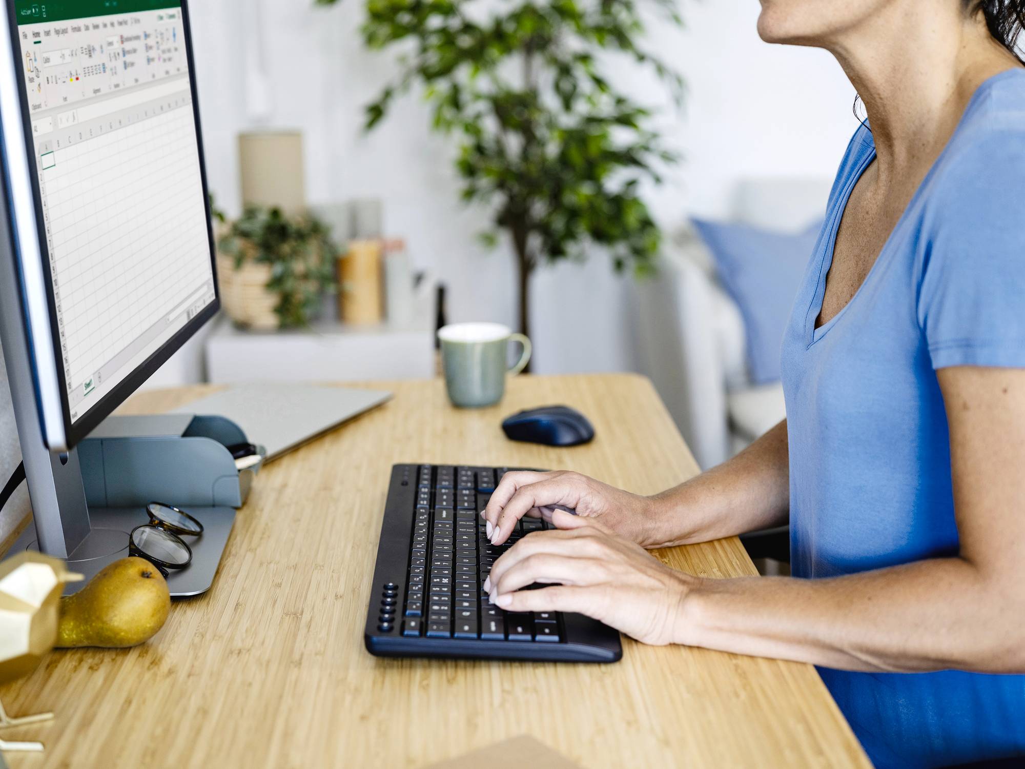 Eine Person tippt auf einer Tastatur vor einem Monitor mit Tabellenkalkulationsprogramm. Im Hintergrund sind Pflanzen und ein Becher zu sehen.