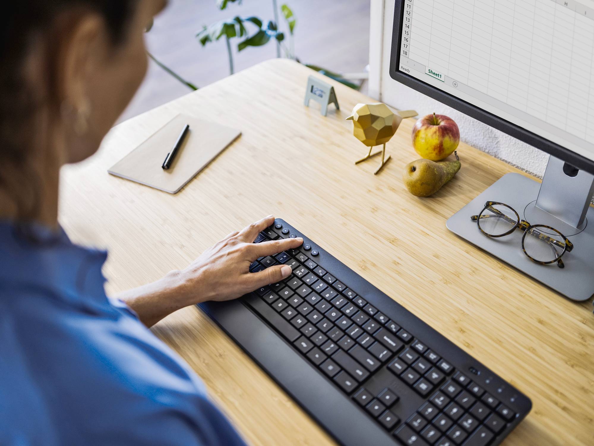 Person arbeitet an einem Schreibtisch mit Tastatur und Monitor, auf dem ein Spreadsheet geöffnet ist. Tisch hat Obst und Brille.
