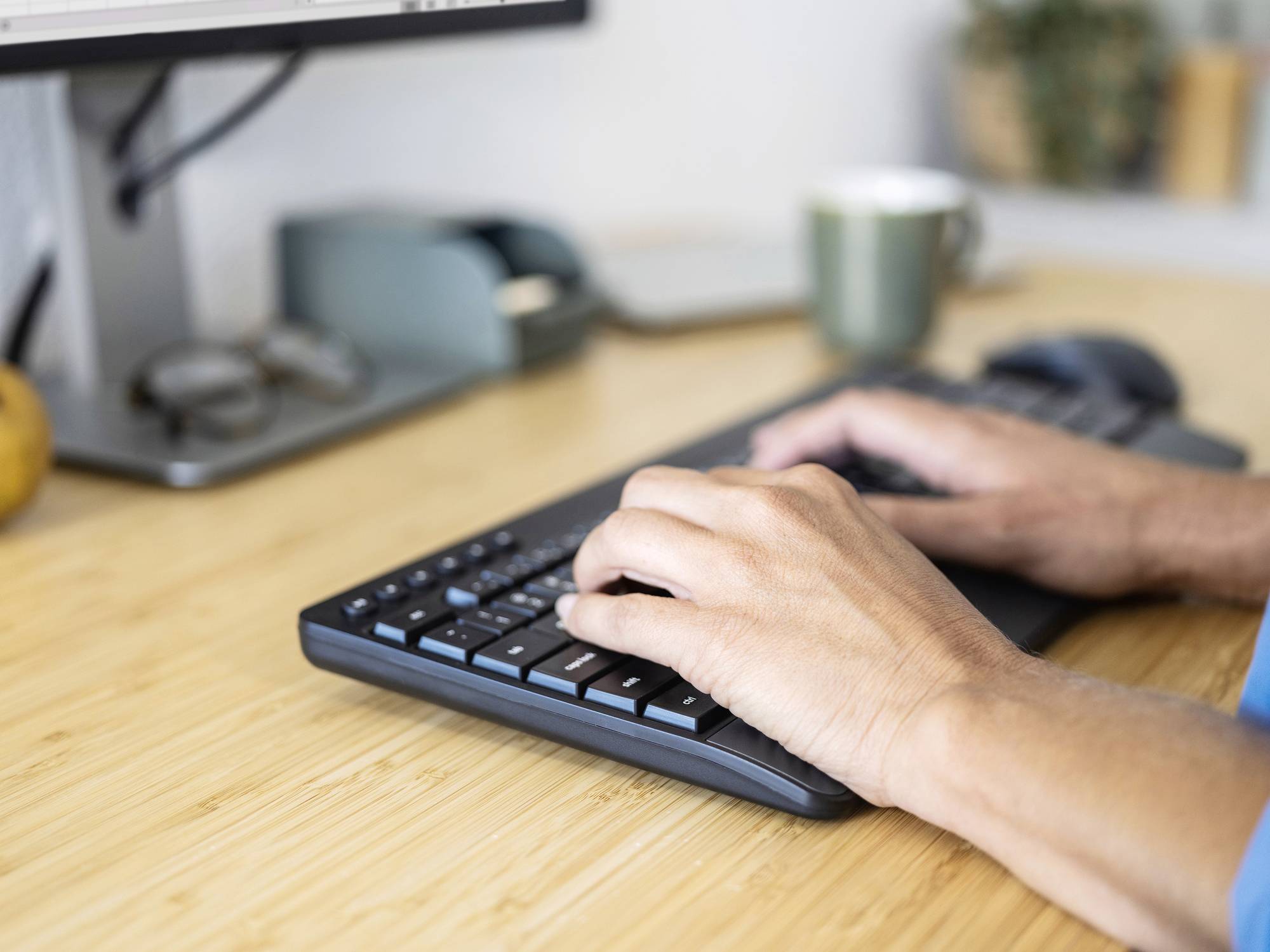 Eine Person tippt auf einer Tastatur an einem Schreibtisch. Im Hintergrund sind ein Monitor, eine Tasse und Pflanzen zu sehen.