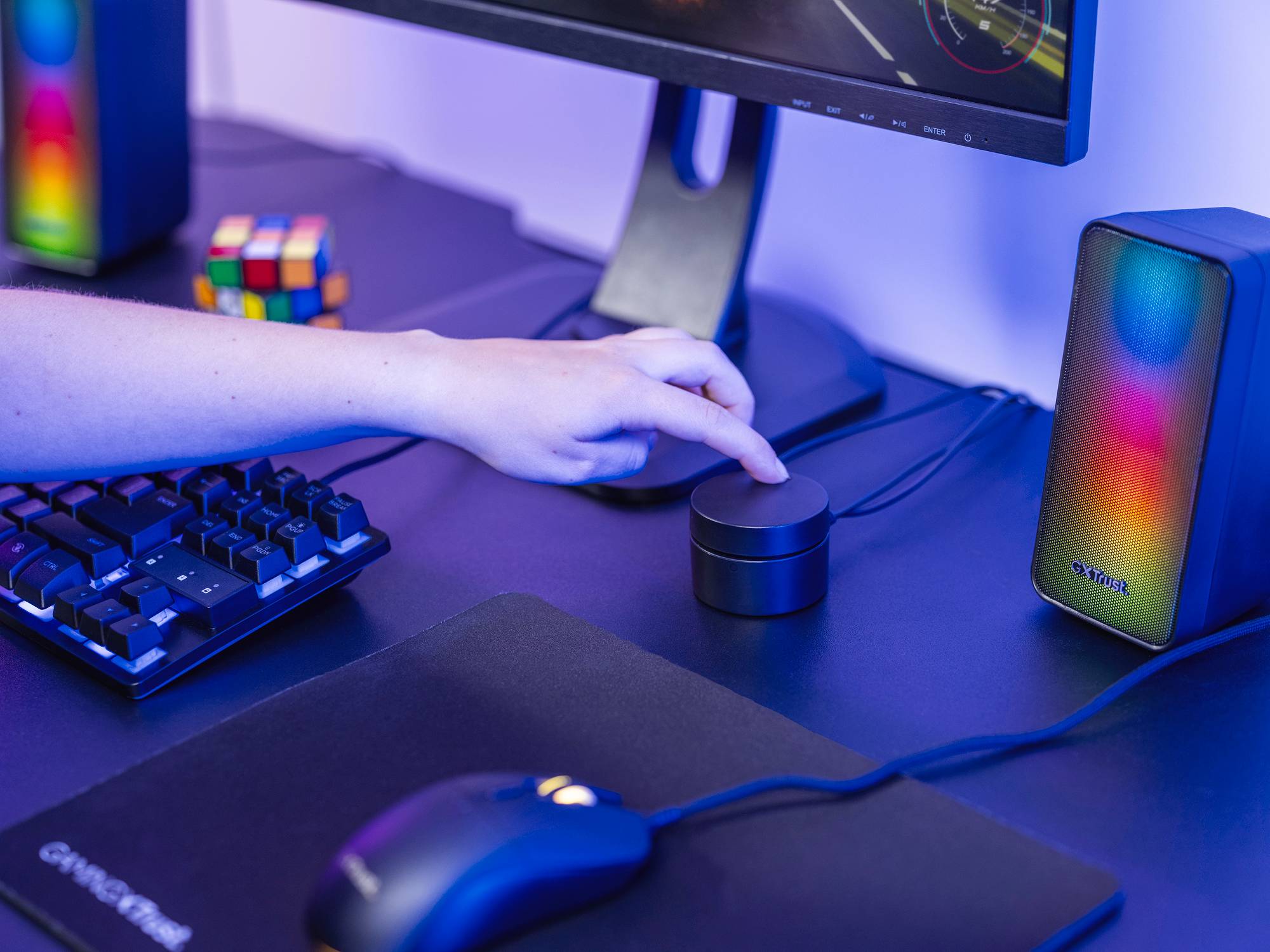 Eine Hand berührt einen runden Lautsprecherregler auf einem Computertisch mit Tastatur, Monitor und buntem Lautsprecher.