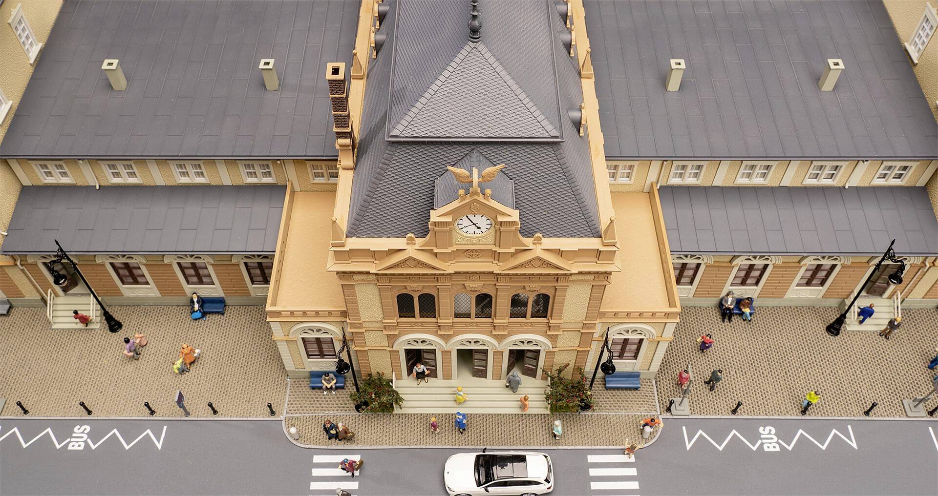 Luftaufnahme eines Miniatur-Bahnhofs mit Uhrturm, umgeben von Menschen und einem vorbeifahrenden Auto auf der Straße im Vordergrund.