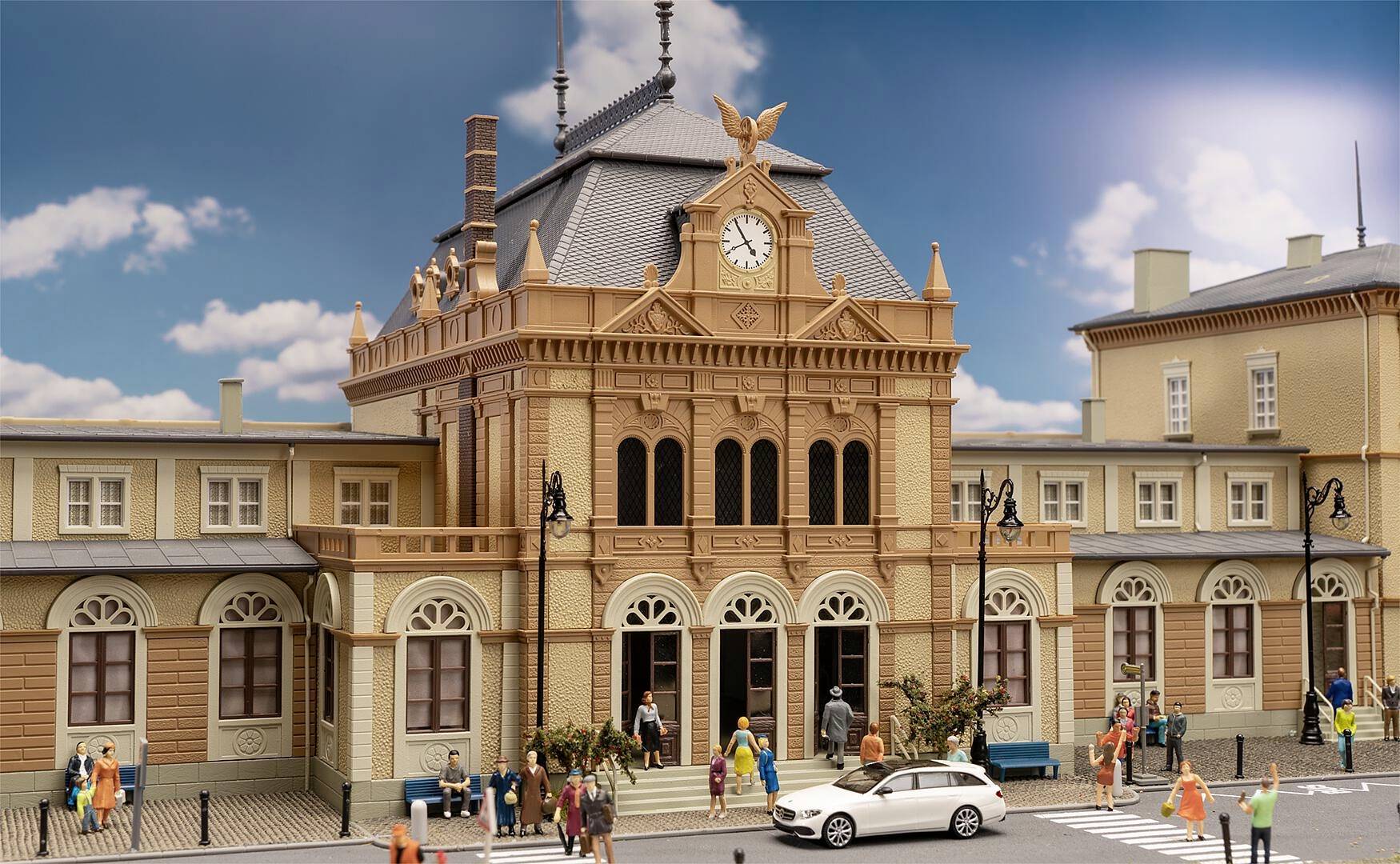 Ein Modell eines historischen Bahnhofsgebäudes mit Uhr und Figuren, die am Eingang verweilen. Im Vordergrund ein weißes Auto.