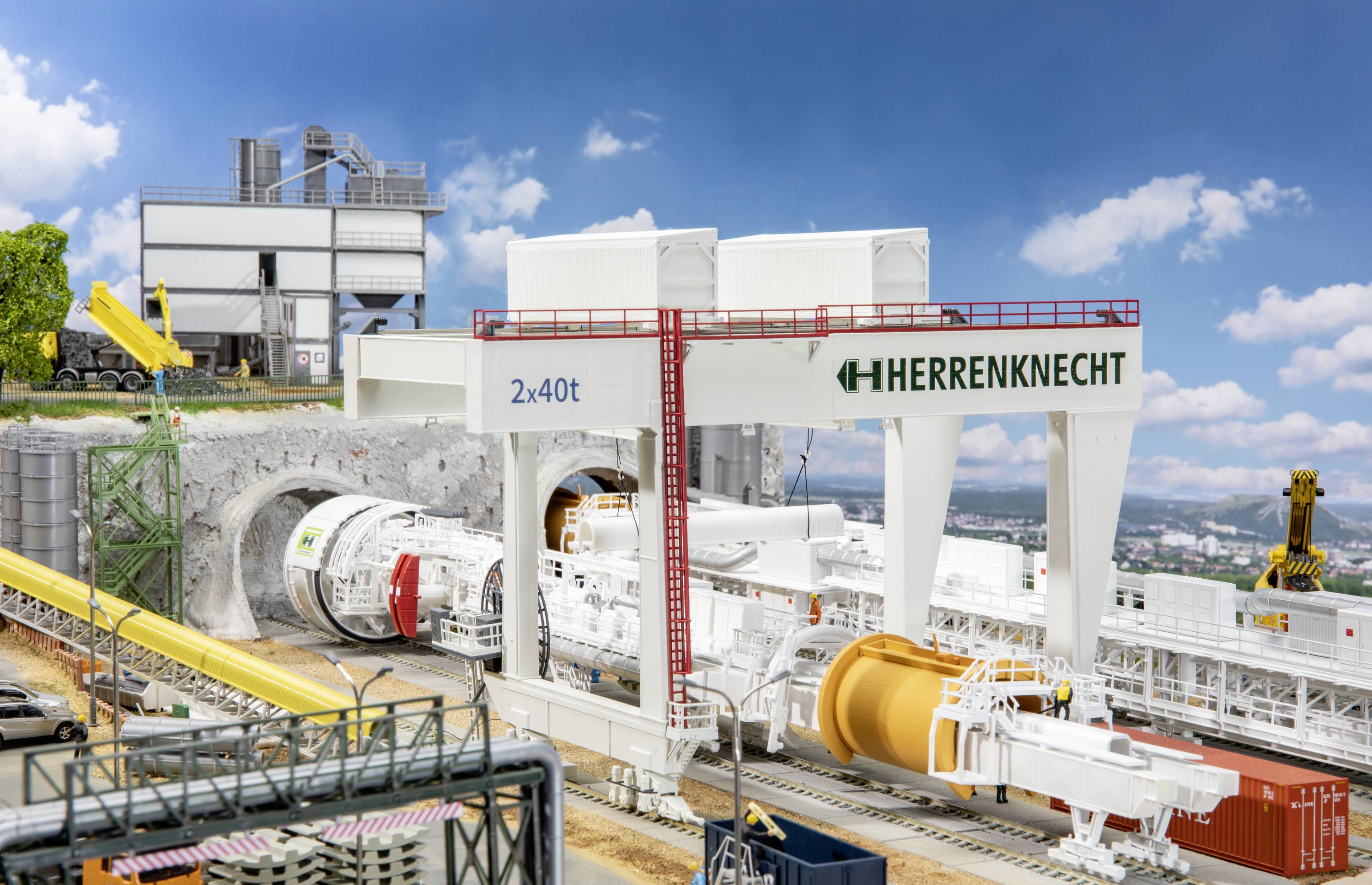 Tunnelbohrmaschine im Einsatz in einer Baustelle; Gebäude und Kräne im Hintergrund; blauer Himmel mit Wolken; 'HERRENKNECHT' Schriftzug sichtbar.
