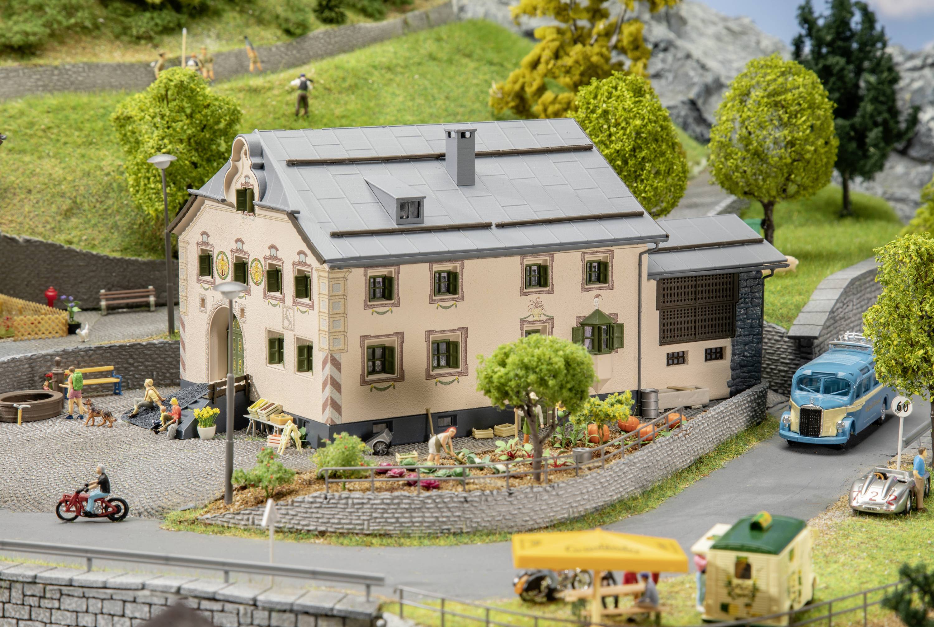 Ein Miniaturdorf mit einem großen rosa Haus in der Mitte, umgeben von Bäumen, Menschen, einem blauen Bus und zwei kleinen Fahrzeugen.