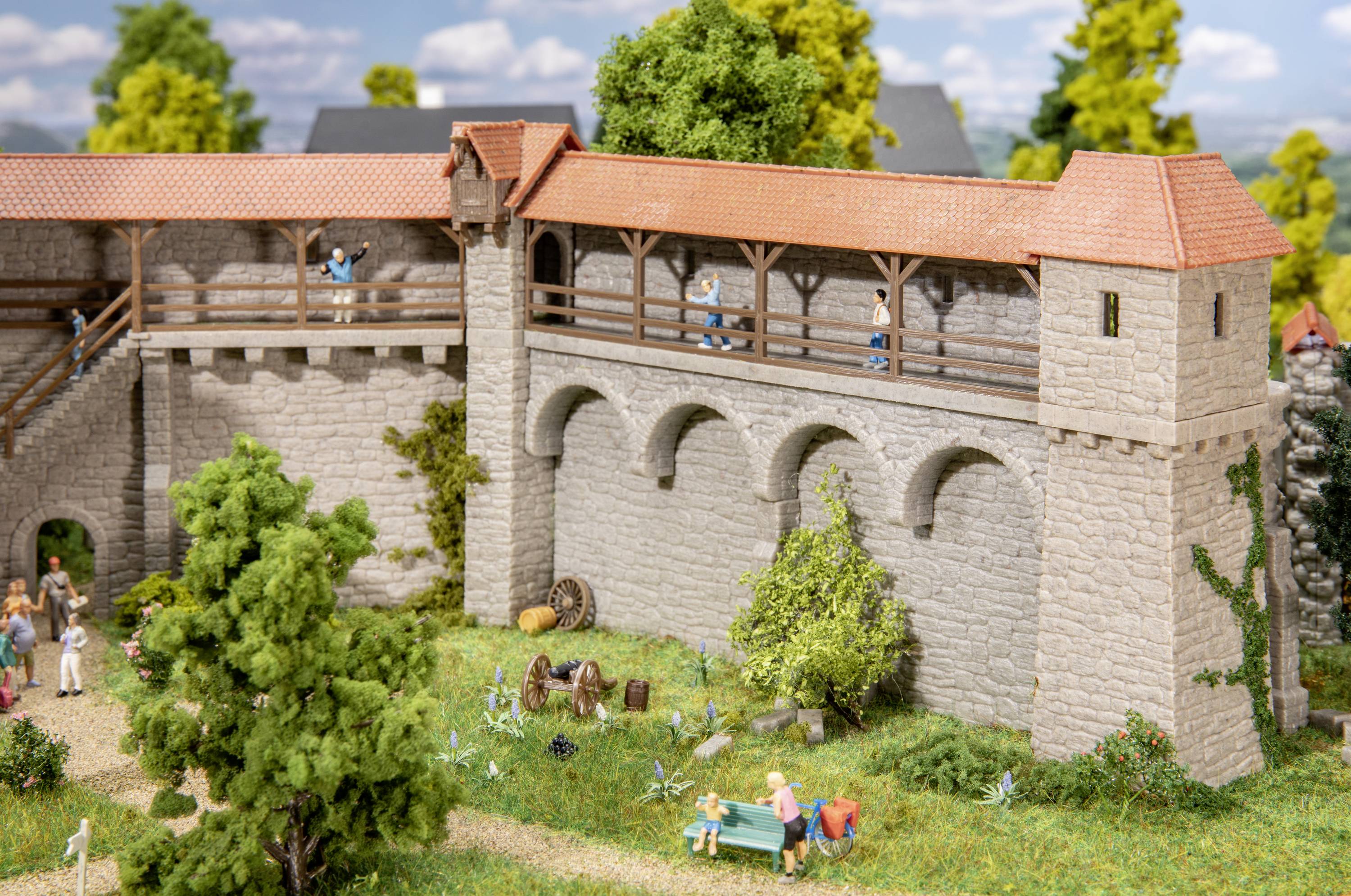 Miniaturmodell einer mittelalterlichen Burgmauer mit Figuren, die spazieren gehen und auf einer Bank sitzen. Bäume und Pflanzen im Vordergrund.