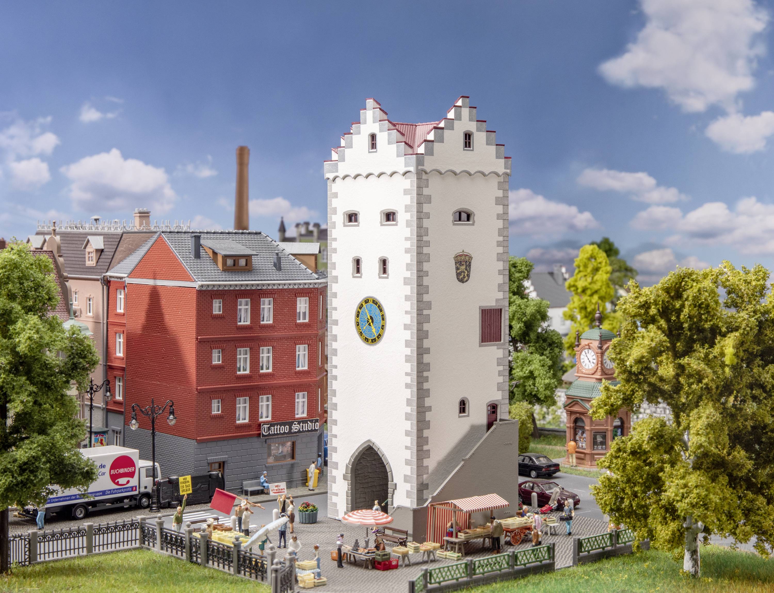 Ein Modell eines mittelalterlichen Stadtturms umgeben von Gebäuden, Bäumen und einem kleinen Markt unter blauem Himmel.