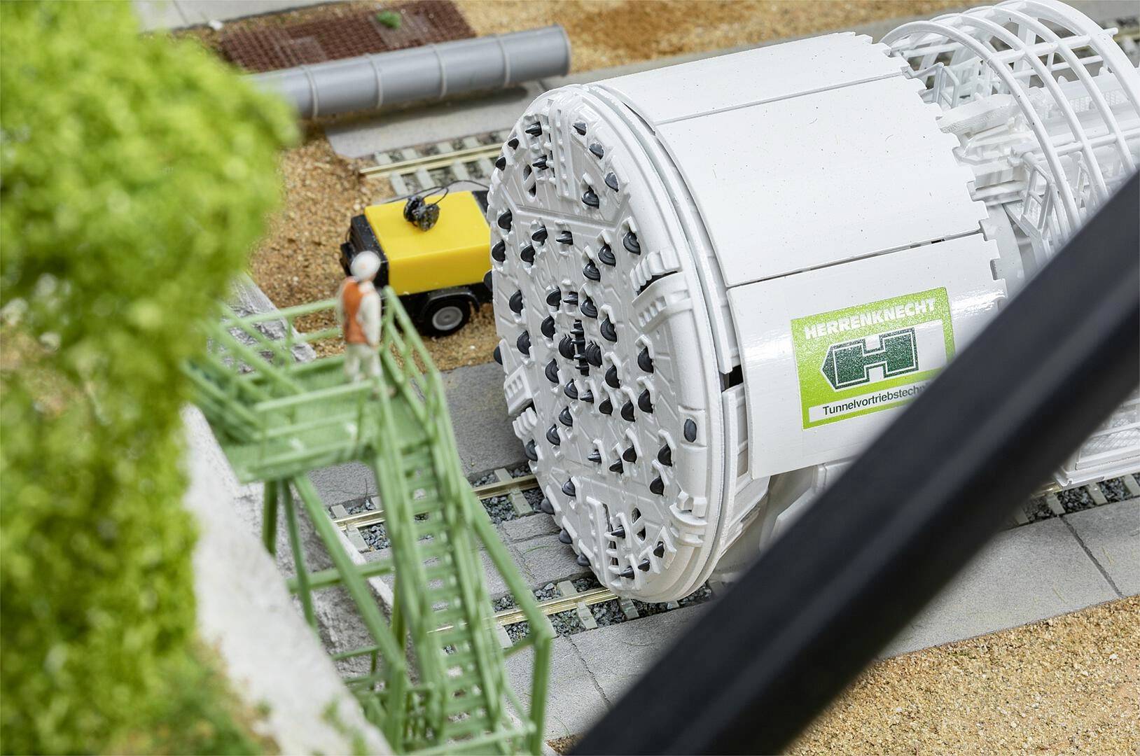 Eine Tunnelbohrmaschine wird nahe Schienen und Grünanlagen installiert. Ein Arbeiter steht auf einem Gerüst und beobachtet den Vorgang.