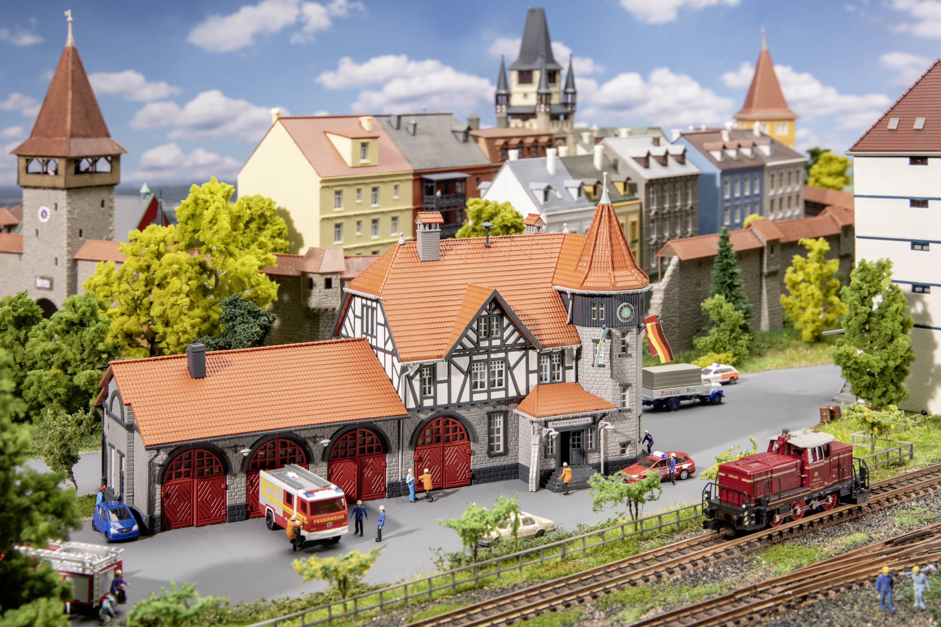 Eine Modelleisenbahnszene zeigt einen Bahnhof mit rotem Ziegeldach, umgeben von kleinen Figuren, Fahrzeugen und einer vorbeifahrenden Lokomotive.