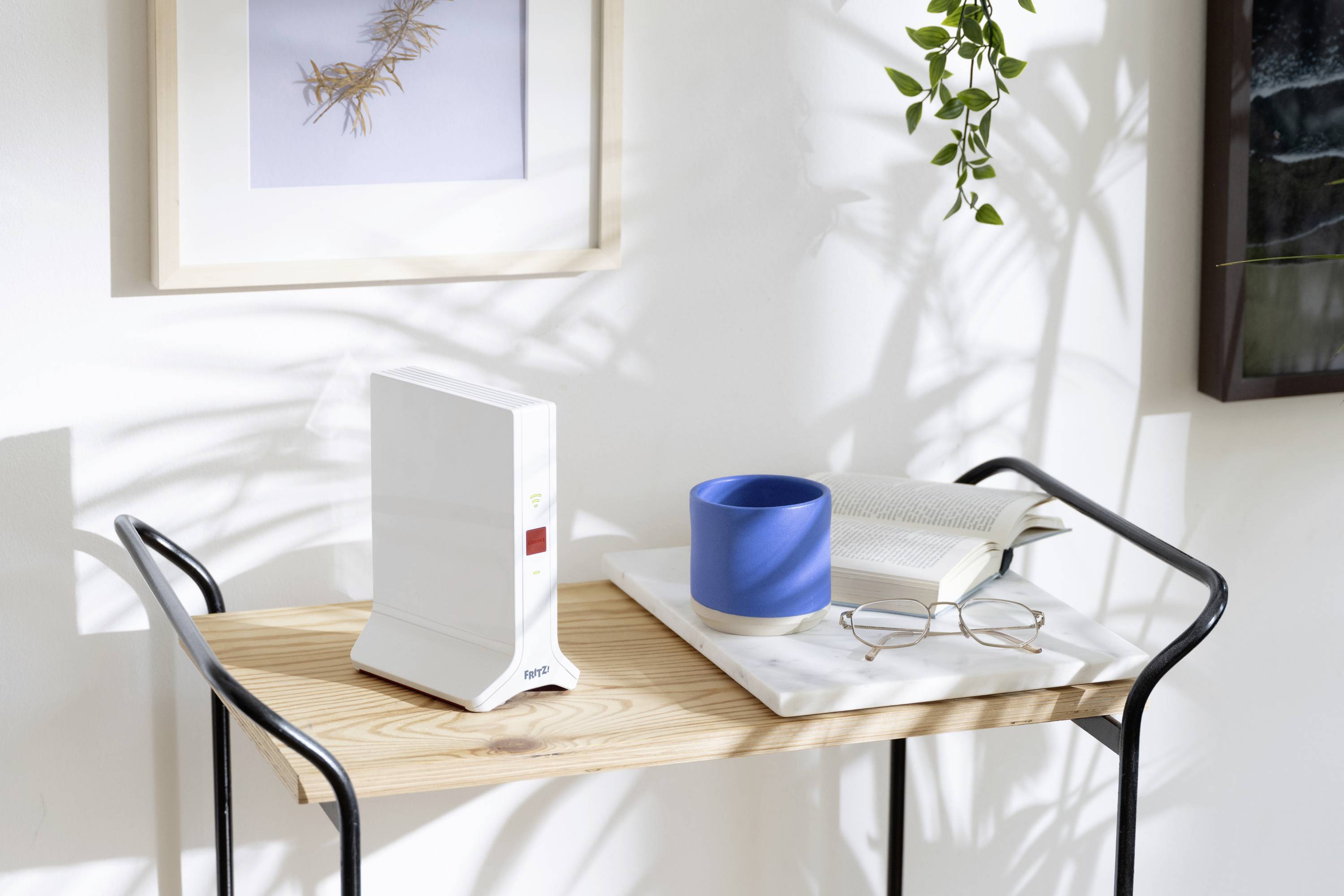 Ein Router auf einem Holztisch neben blauem Becher, Buch und Brille, umgeben von Pflanzen im Sonnenlicht, schafft eine gemütliche Atmosphäre.