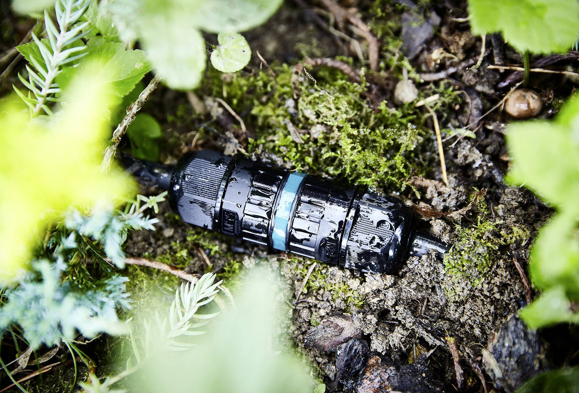 Eine schwarze Steckverbindung mit blauem Ring liegt auf feuchtem Boden, umgeben von grünen Blättern und Pflanzen im Garten.