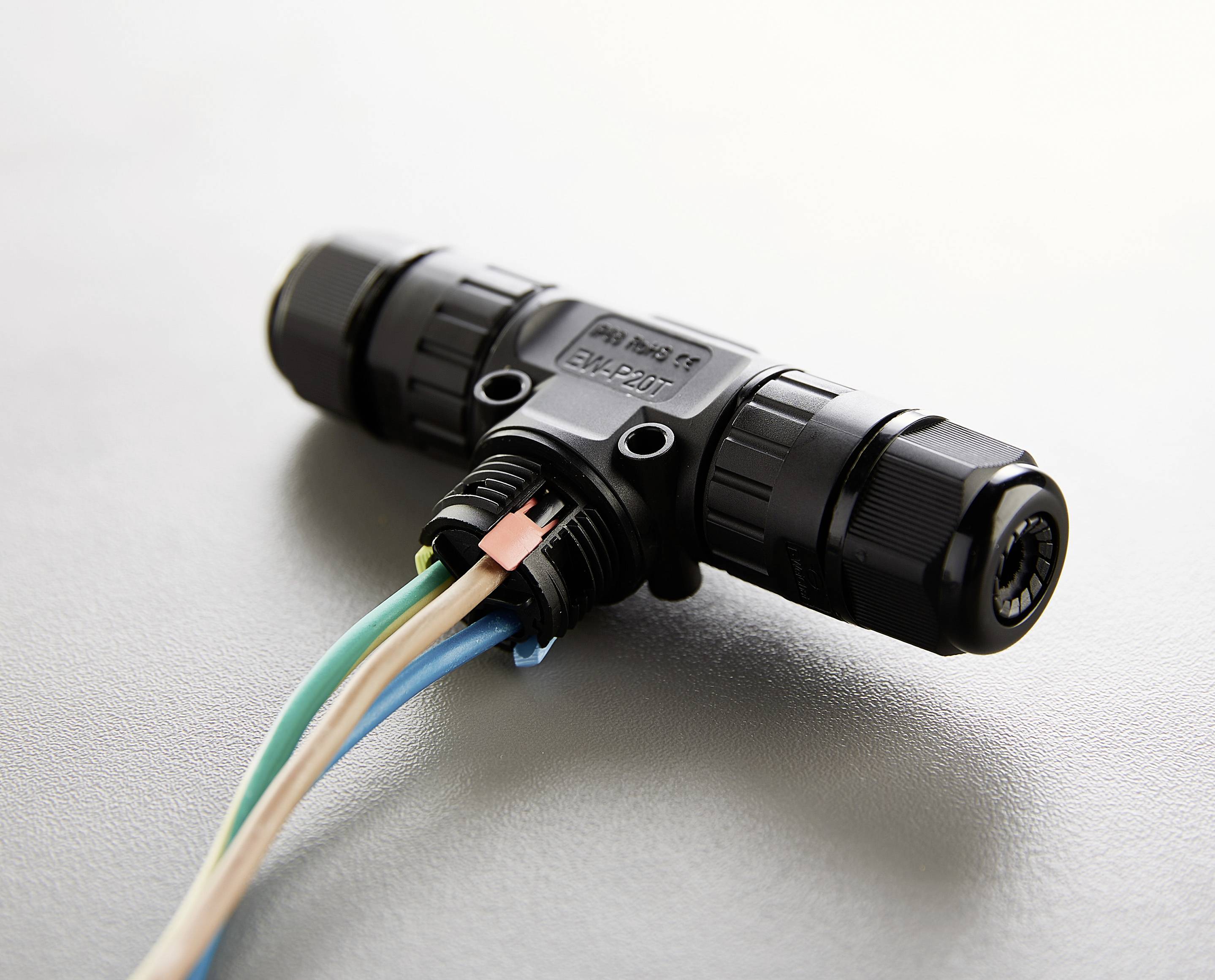 Ein schwarzer Kabelverbinder in T-Form mit mehreren farbigen Drähten, auf grauer Oberfläche liegend.