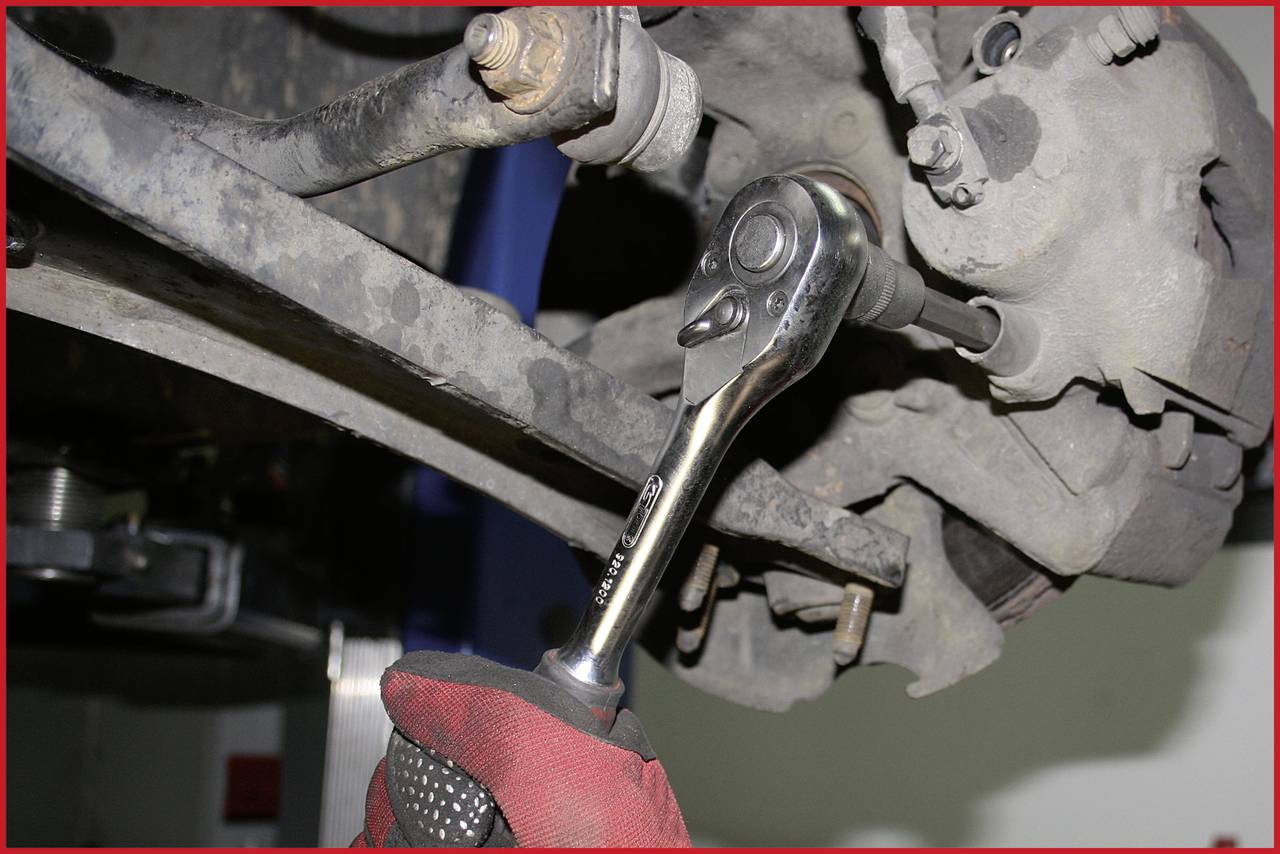 Person mit Handschuh repariert Autoteil mit Ratsche unter Fahrzeug.