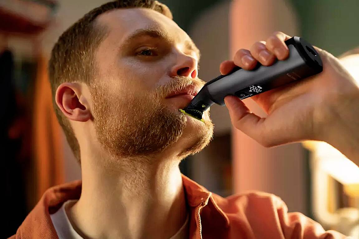 Ein Mann rasiert sich den Bart mit einem elektrischen Rasierer. Er trägt ein orangefarbenes Hemd. Die Umgebung ist weich beleuchtet.