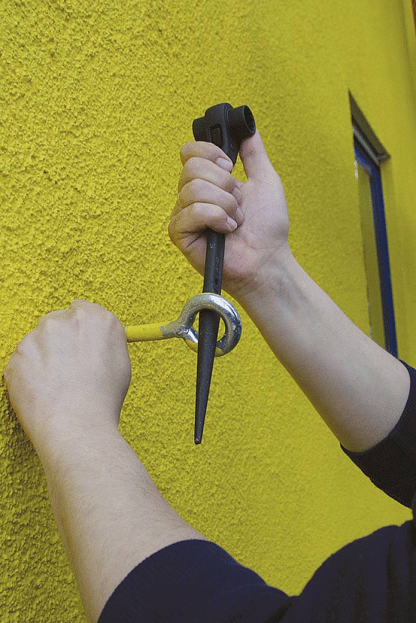 Eine Person benutzt einen Schraubenschlüssel, um einen Ringanker an einer gelben Wand zu befestigen.