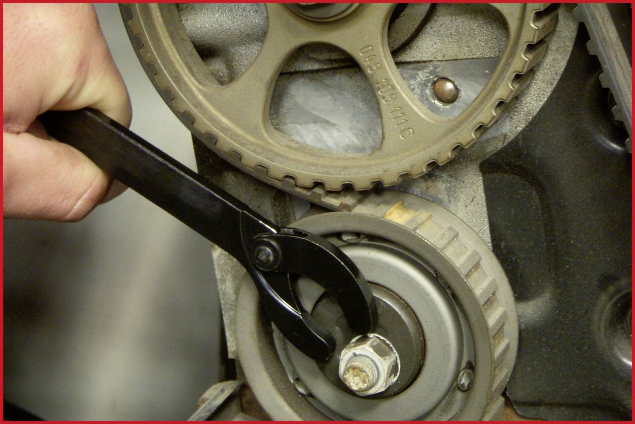 Eine Person verwendet einen schwarzen Schraubenschlüssel, um an einem Motorriemen zu arbeiten. Details der Zahnräder sind sichtbar.