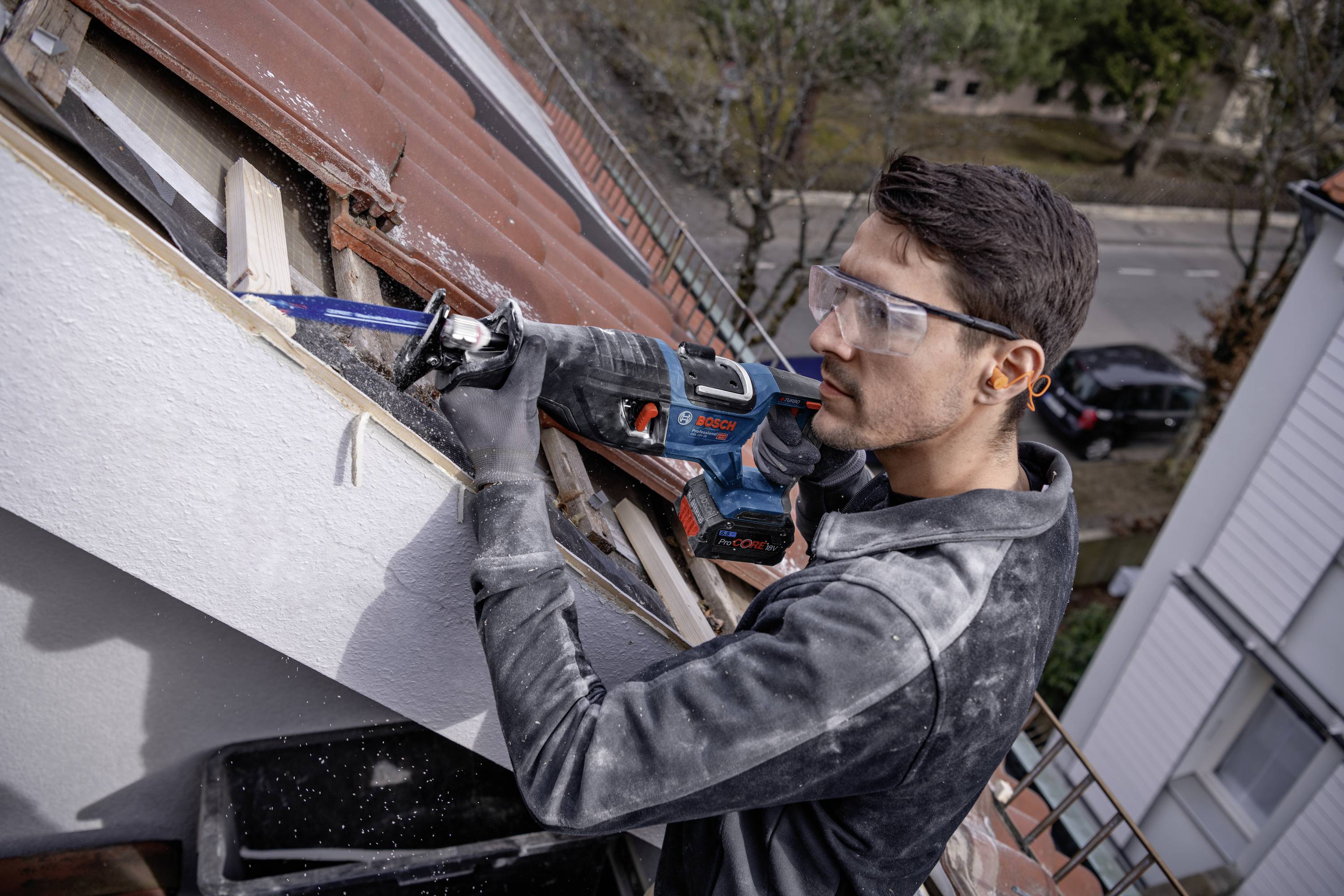 Ein Mann mit Schutzbrille verwendet ein elektrisches Sägetool, um ein Holzbrett an einem Dach zu schneiden. Im Hintergrund sind Bäume und eine Straße zu sehen.