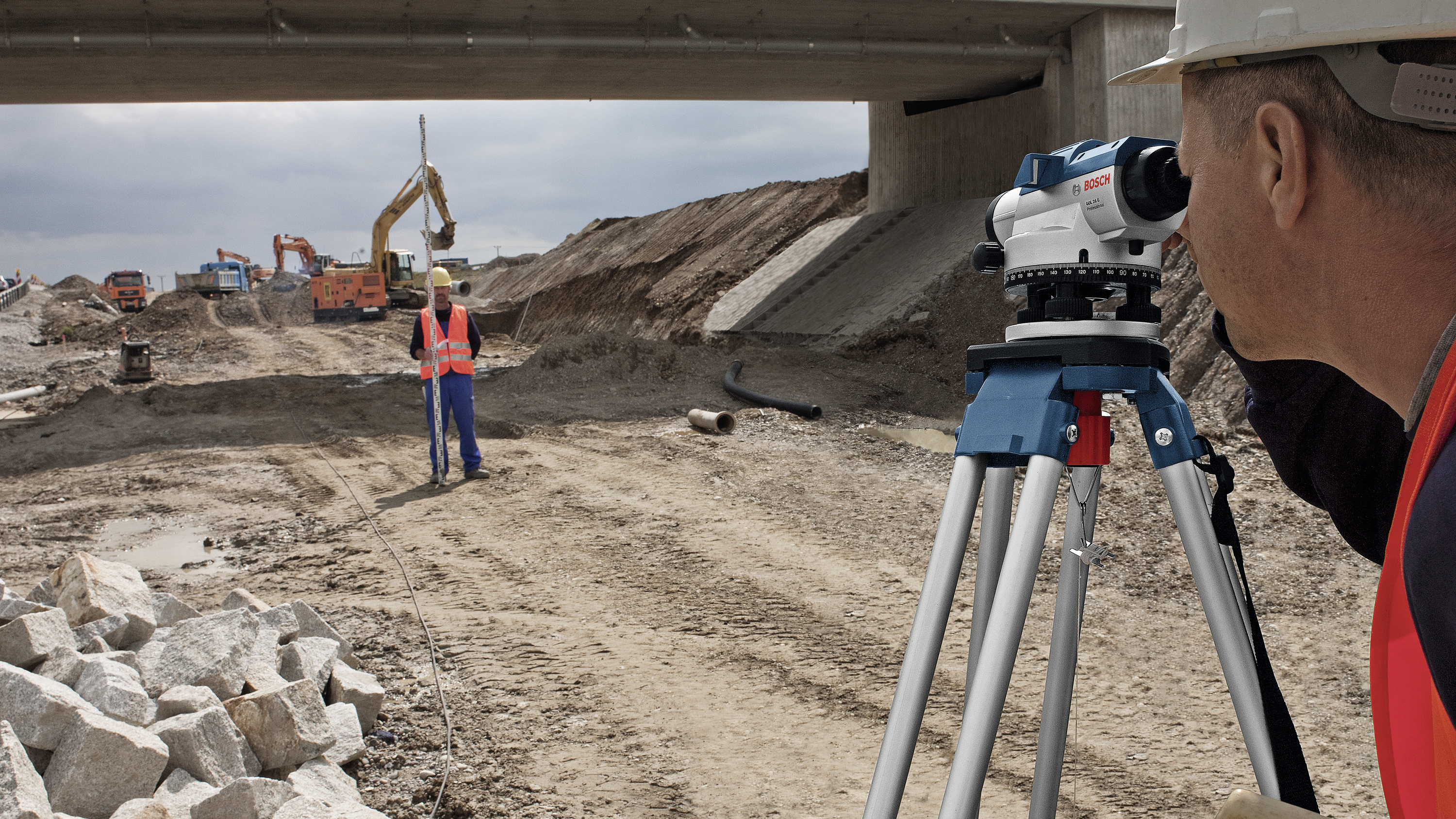 Ein Vermessungsingenieur verwendet ein Theodolit, um eine Baustelle unter einer Brücke zu vermessen. Im Hintergrund arbeiten Bagger.