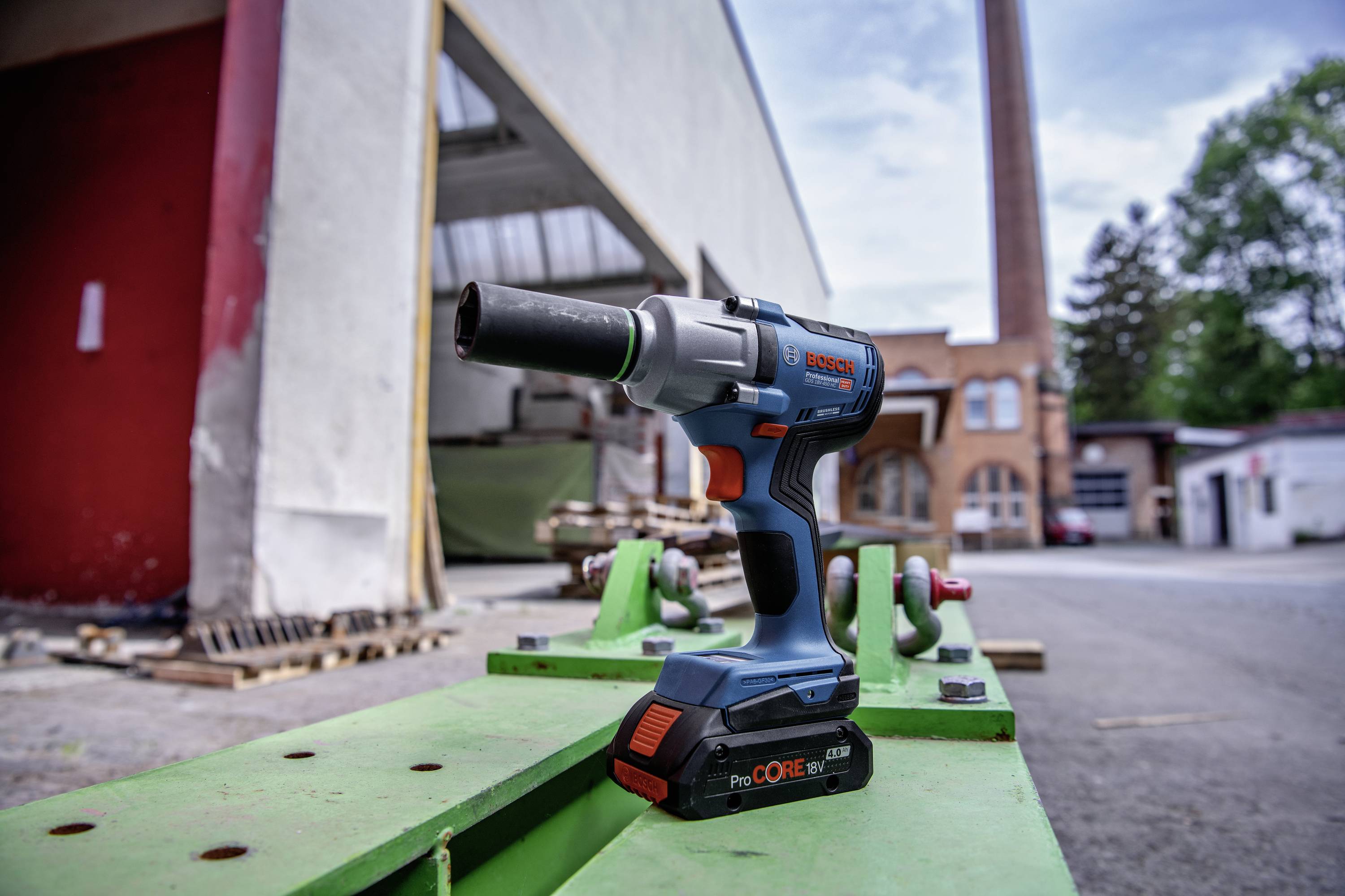 Ein blau-oranger Akkuschrauber steht auf einem grünen Tisch, im Hintergrund sind eine Fabrikhalle und ein Schornstein sichtbar.