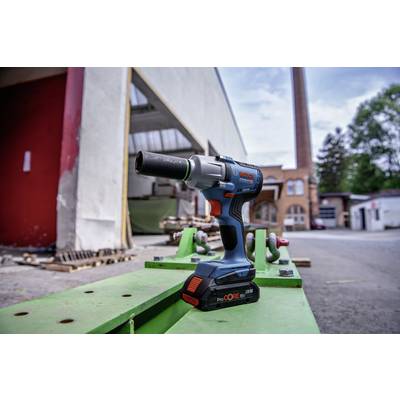 Ein blau-oranger Akkuschrauber steht auf einem grünen Tisch, im Hintergrund sind eine Fabrikhalle und ein Schornstein sichtbar.