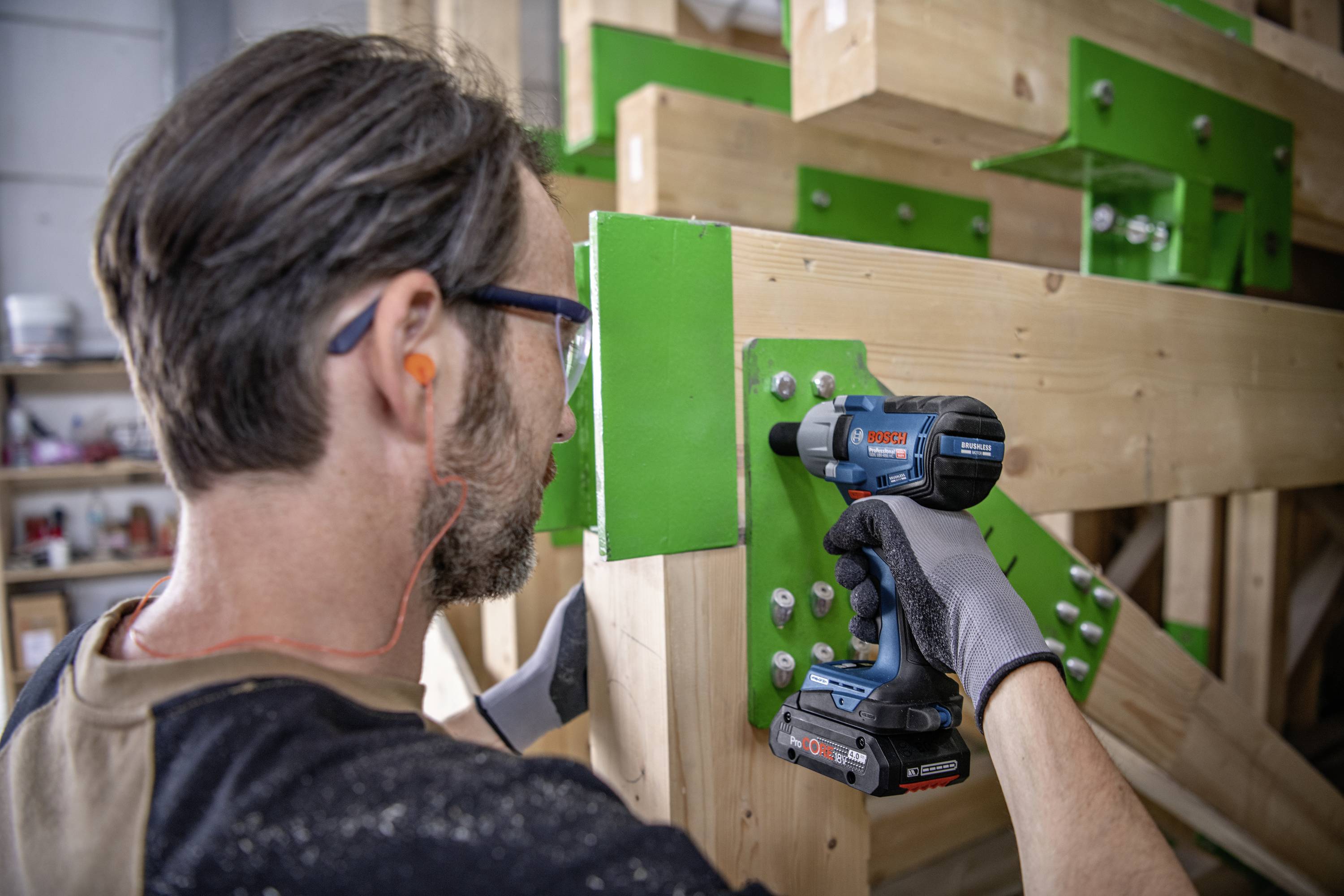 Ein Handwerker befestigt Holzträger mit einem Akku-Bohrschrauber. Er trägt Gehörschutz und Arbeitshandschuhe, arbeitet an einer Holzkonstruktion.